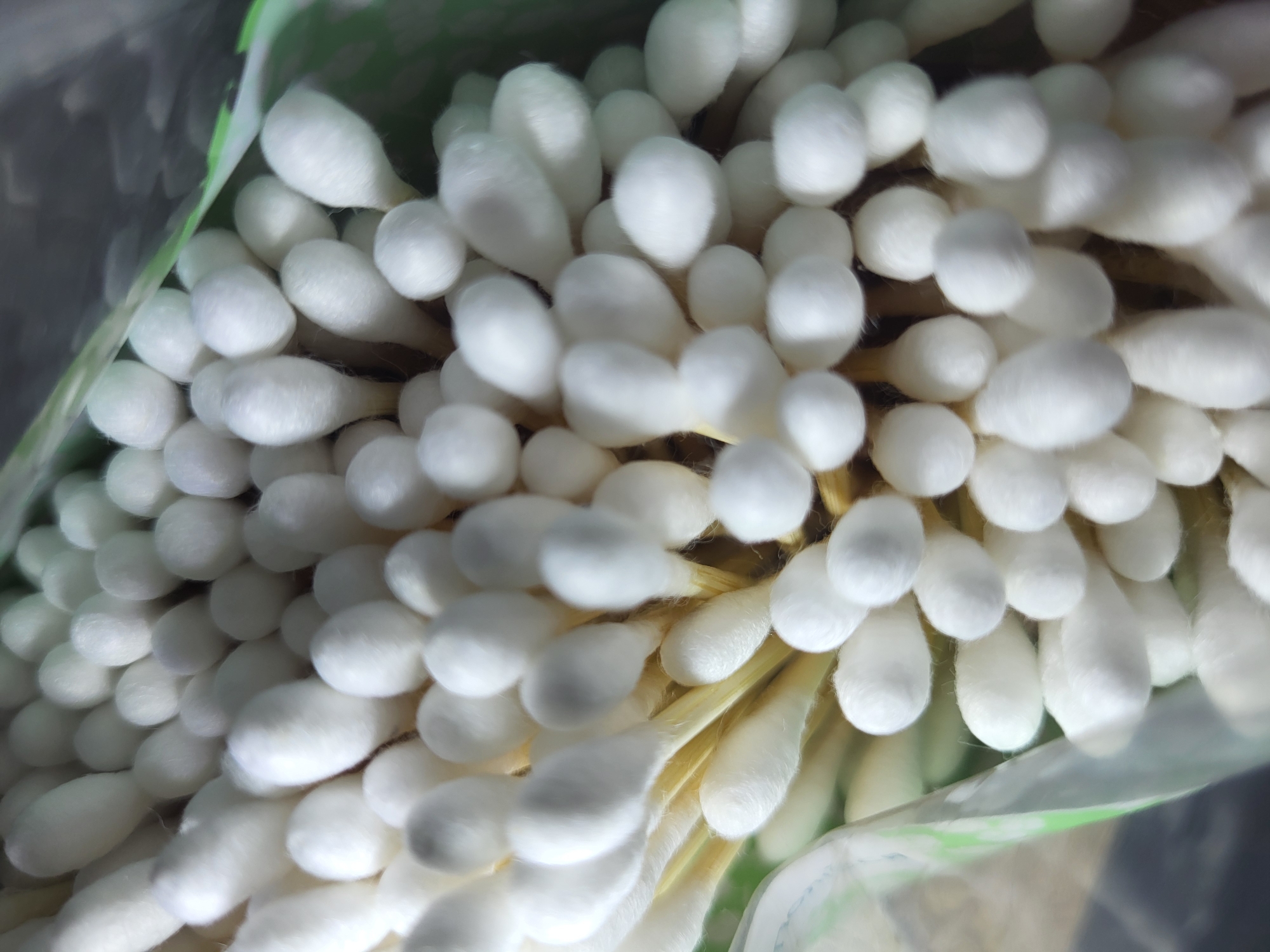 A close-up, overhead shot shows a bundle of white cotton swabs with yellowish paper sticks, partially enclosed in clear plastic packaging. The packaging is shiny and reflective, with green trim visible along the top edge. The cotton swabs are densely packed and fill the frame, with some appearing blurred due to shallow depth of field. The background is dark and indistinct, suggesting an indoor setting or the bottom of a bag. The image does not depict any specific location in the city of Horishni Plavni, Ukraine.