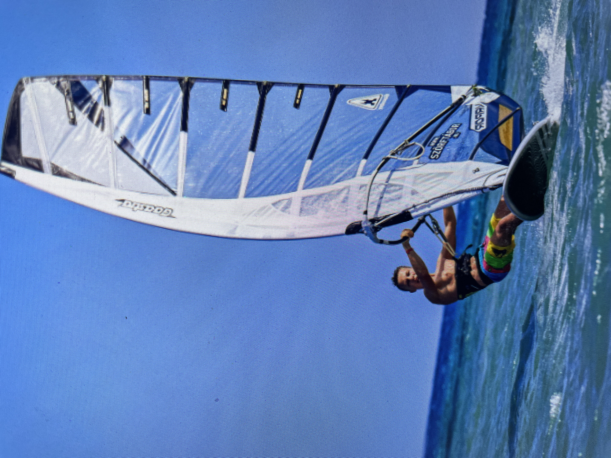 A windsurfer is in action. The subject is a male wearing colorful shorts and no shirt. He is holding the boom of a windsurf sail. The sail is white and blue. The water is visible. The sky is blue and clear. There is no visual confirmation the image was taken in Bacong, Philippines.