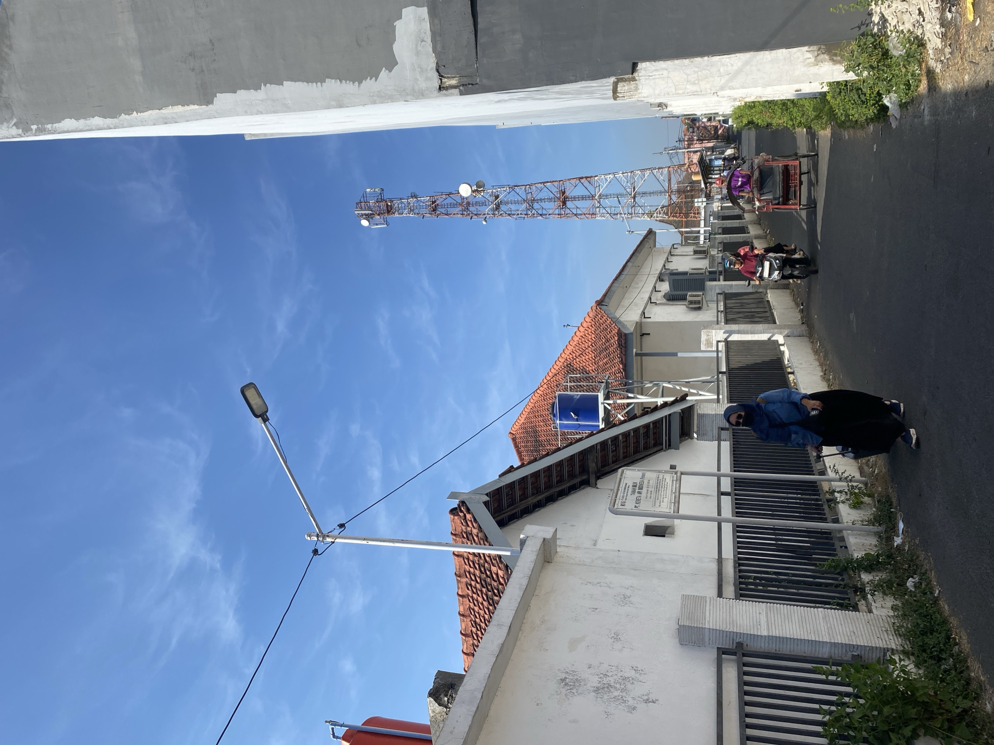 The image depicts a street scene in Kediri City, Indonesia, characterized by a bright blue sky with sparse white clouds. A tall telecommunications tower with red and white lattice structure stands prominently in the background. On the left side of the street, there are white buildings with red tile roofs. A street lamp is visible overhead, supported by a pole. A woman wearing a blue hijab and denim jacket, along with a black skirt and sunglasses, walks along the pavement pulling a suitcase on wheels. She is on the left side of the road. Further down the street, a man rides a motorcycle, and behind him, a person is operating a pedal-powered rickshaw. The road is paved and appears to be narrow, with vegetation growing along the sides of the buildings and the road. There are also some water tanks visible on the roofs of the buildings. A sign is mounted on a pole in front of one of the buildings. The overall environment suggests a residential or mixed-use area within a city.