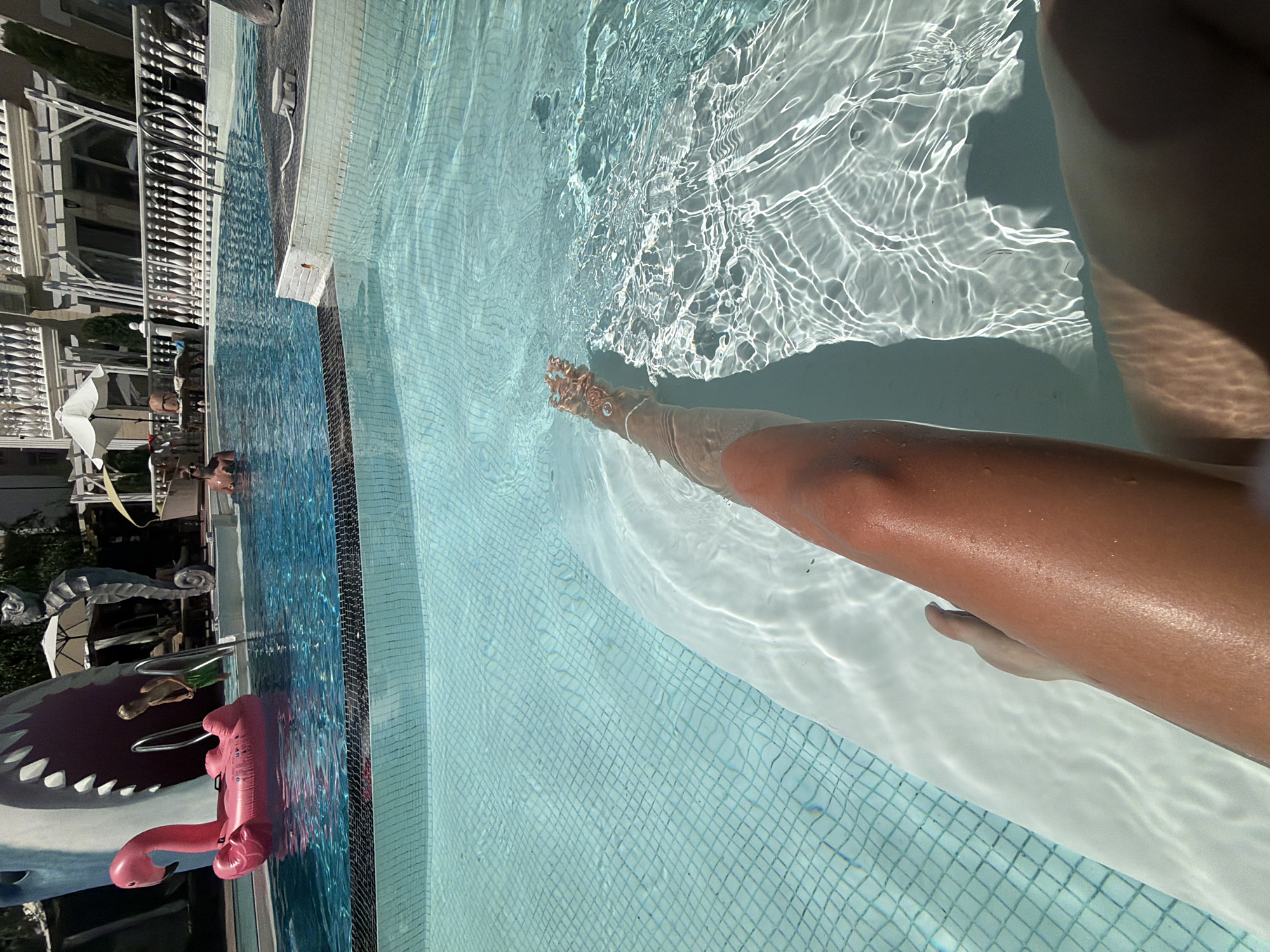 This is a first-person perspective media file of someone's legs submerged in a swimming pool. The water is clear and reflects sunlight, creating rippling patterns on the tiled bottom. The pool appears to be outdoors, with a white railing and some greenery visible in the background, suggesting a sunny day. A pink inflatable flamingo and a large inflatable shark are floating in the water, adding a playful element to the scene. The person's skin is tanned, and they have a silver anklet on their right foot. The overall mood is relaxed and summery. The location context is Nesebar, Bulgaria, which is a coastal town known for its historical sites and beaches.