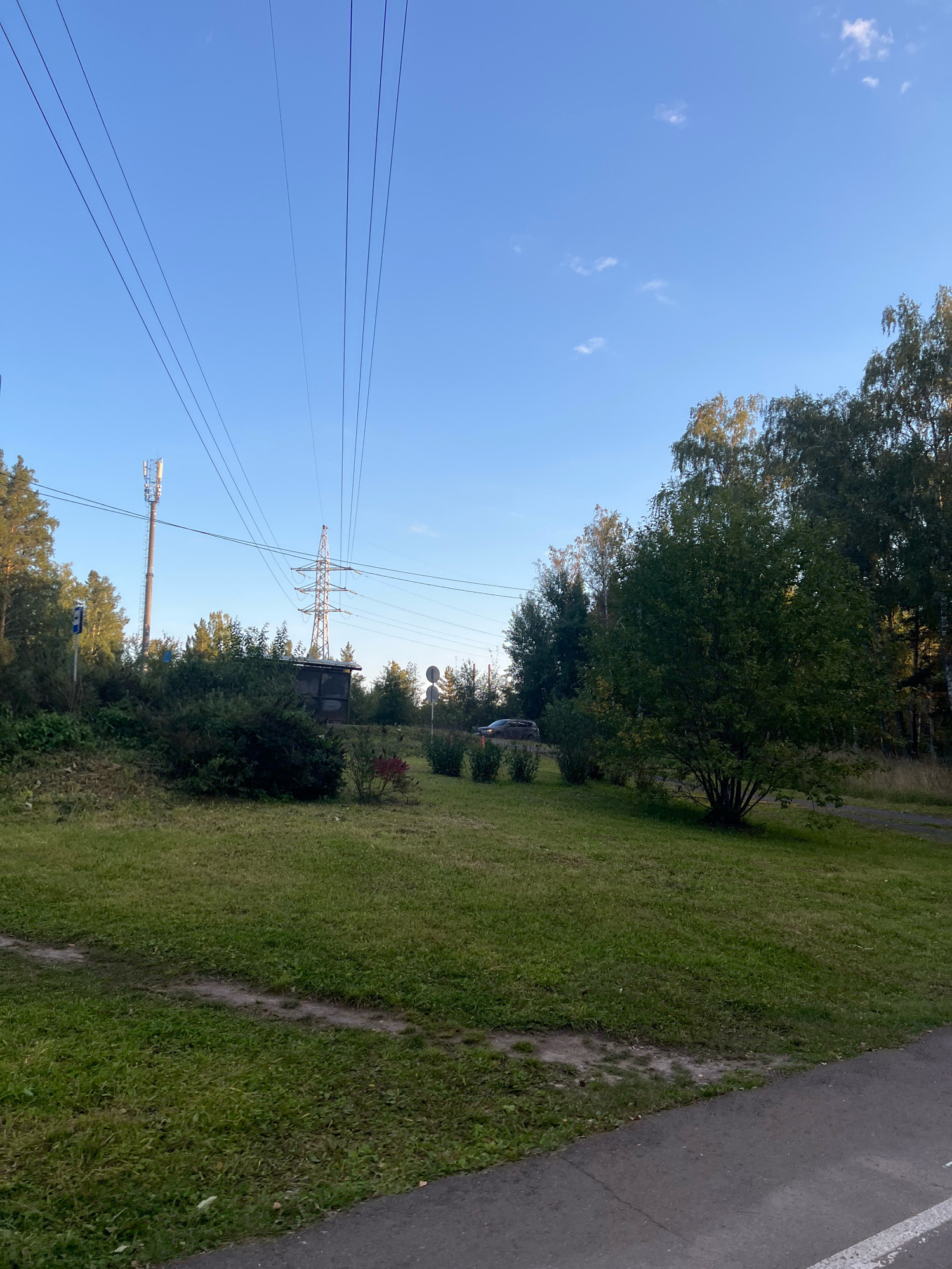 The image depicts a roadside scene with a clear blue sky and scattered clouds. In the foreground, a paved surface, likely a sidewalk or bike path, runs along the bottom right edge. Behind this, a grassy area with some bare patches and faint tracks leads to a cluster of green bushes and trees.
Dominating the mid-ground are several elements of infrastructure. A tall, slender cellular tower with antennas is visible on the left, partially obscured by trees. Further to the right, a large, lattice-style electrical pylon stands, carrying several high-voltage power lines that stretch across the sky. A dark, rectangular structure, possibly a small shed or utility building, is situated behind the bushes near the pylon.
In the distance, a dark-colored car is driving on a road that curves through a wooded area. The trees are predominantly deciduous with green foliage, suggesting a late summer or early autumn setting. The overall environment appears to be a semi-urban or suburban area adjacent to a natural landscape. The presence of power lines and a cellular tower indicates developed infrastructure within a somewhat naturalistic setting.
Given the information that the image is from Krasnoyarsk, Russia, this scene could be representative of the outskirts of the city or areas where urban development meets forested regions. Such a view is common in Russia, where large green spaces often coexist with essential utility infrastructure.