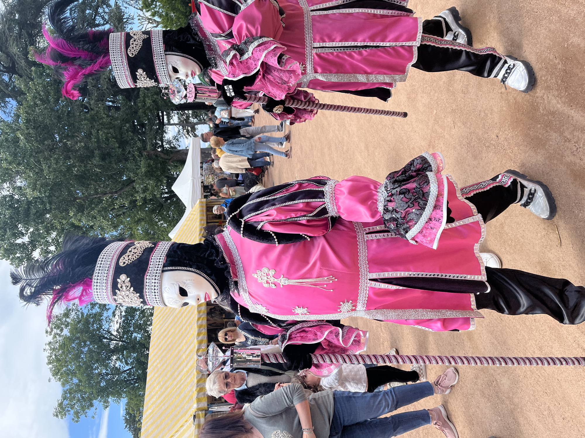 The image depicts two individuals dressed in elaborate, historical-style costumes in what appears to be a public outdoor setting. The costumes are predominantly pink and black, with silver trim and ornate details. The individuals are wearing full-face white masks and tall, feathered hats. Each is holding a long, spiraled baton.
The background includes other people, some tents or canopies with yellow and white stripes, and trees. The ground is a light brown color, possibly dirt or gravel. The presence of tents and the crowd suggests an event or festival.
Given the location in Saint-Galmier, France, and the style of the costumes, it is plausible this is related to a local festival, perhaps of historical or cultural significance.