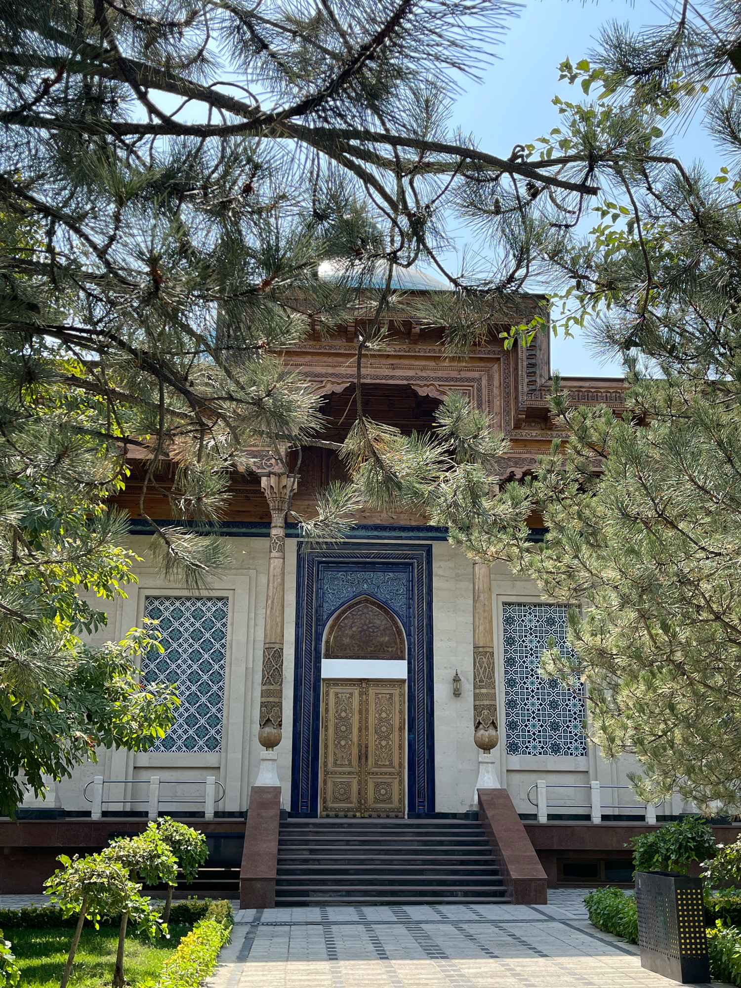 The image captures the front facade of a building, likely a historical or religious site, viewed through the branches of pine trees. The building features intricate wooden carvings above a grand entrance with ornate golden doors. Flanking the entrance are large windows with geometric patterns in blue and white. The facade is constructed of light-colored stone, accented by dark blue tile work around the doorway and windows. A set of dark stairs leads up to the entrance. In the foreground, manicured bushes and trees line a paved walkway, with a black trash receptacle visible on the right. The sky above is clear and blue, indicating daytime. This building is located in Tashkent, Uzbekistan, and is identified as the Minor Mosque.