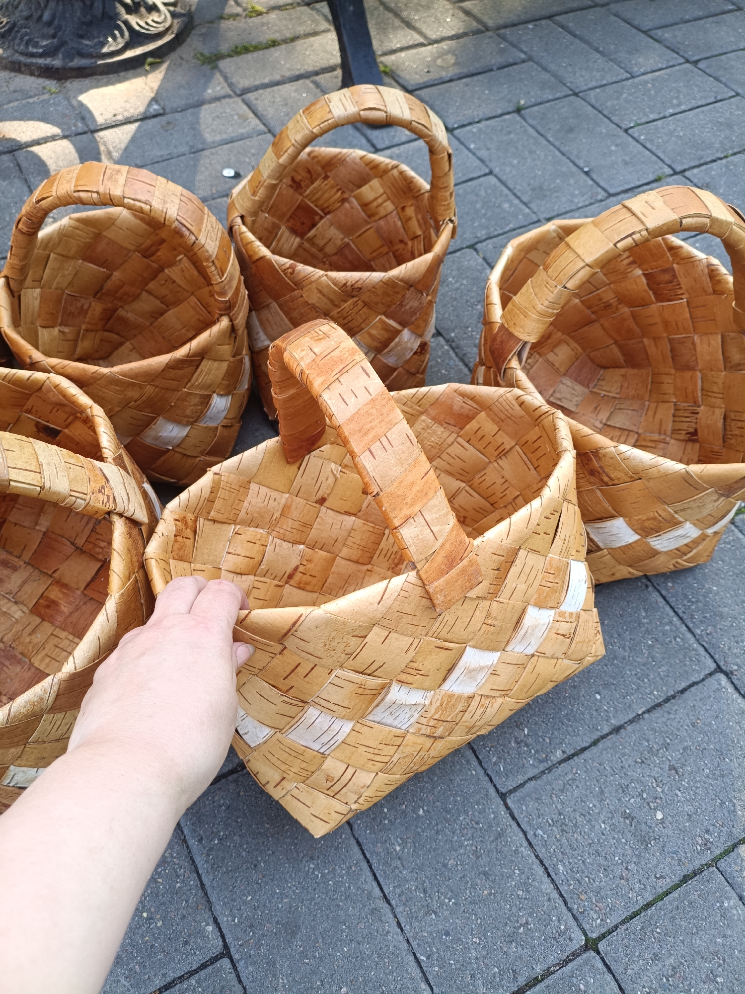 The image shows a collection of five birch bark baskets arranged on a paved surface. The baskets are of various sizes and feature woven patterns with some white accent bands. One hand is reaching into the closest basket. The background consists of dark gray paving stones and a hint of greenery and a dark metal object, possibly a table leg. The scene appears to be outdoors, likely on a sidewalk or patio. The image quality is good, with clear focus on the baskets and the hand. There is no identifiable location information present in the image itself that would indicate it is from Yaroslavl, Russia.