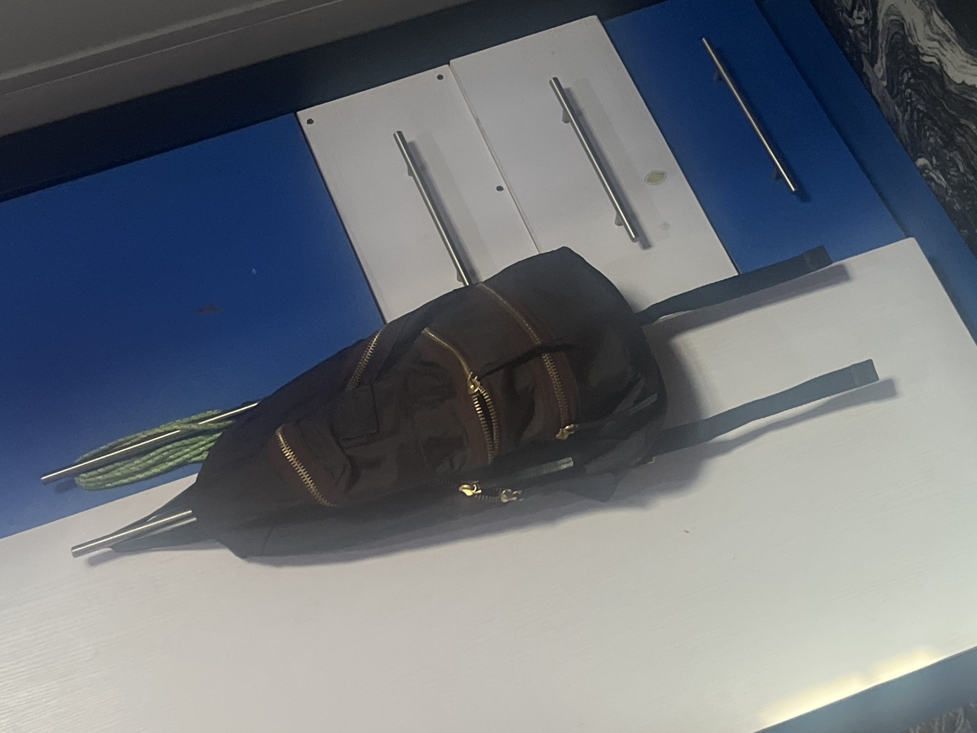 A dark-colored backpack hangs from a hook on a white, textured wall. To the right of the wall is a blue surface with three white drawers, each with a silver handle. The backpack has several zippered pockets with gold accents. Beside the backpack, also hanging from the hook, is a bundle of green cord. The image appears to be taken indoors, likely in a room with furniture. There is no indication of the city of Gura Top, Nigeria.
