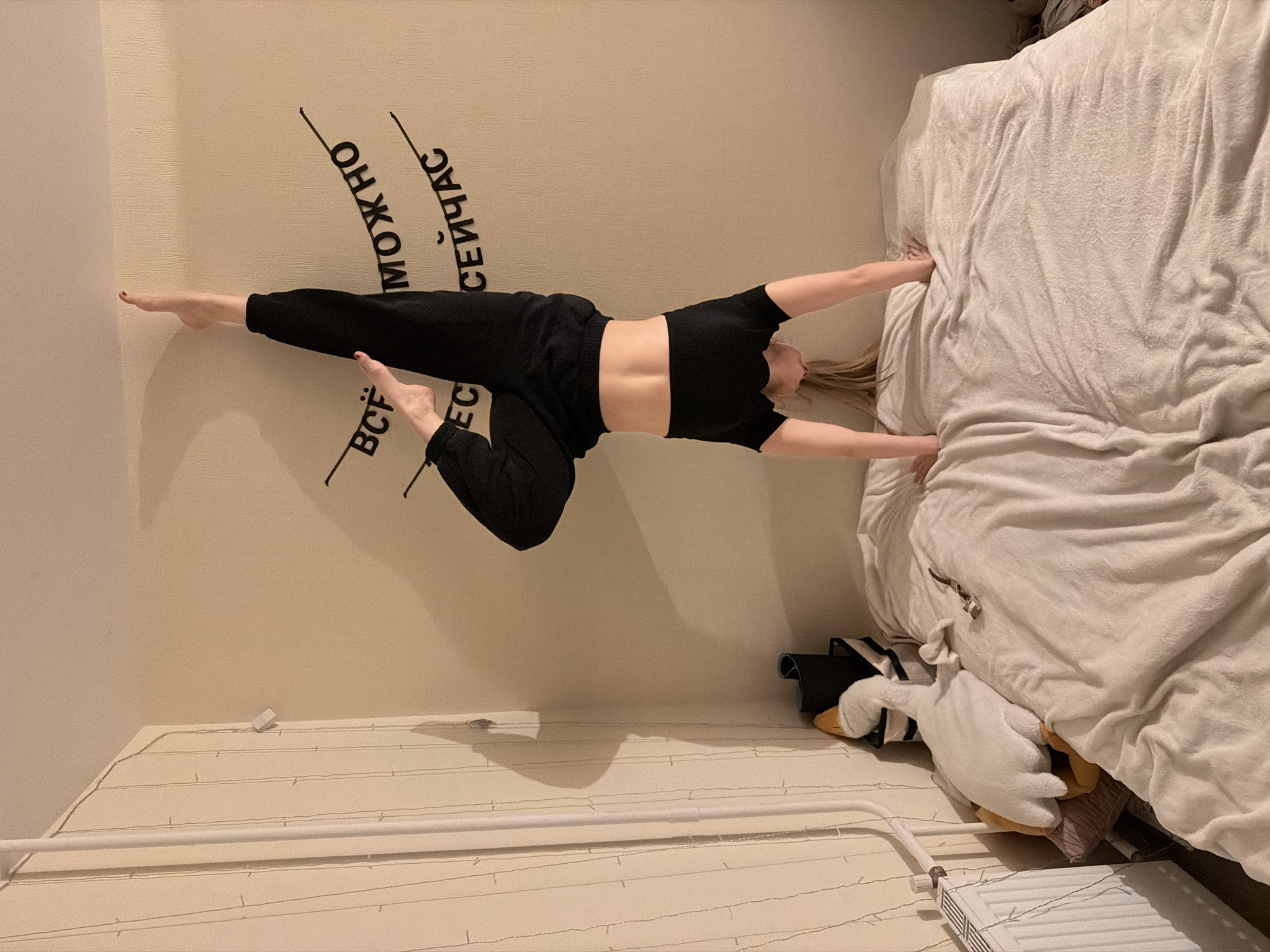 A young woman is captured indoors, performing an acrobatic or yoga pose against a light beige wall. She appears to be suspended horizontally, with her body parallel to a large bed covered with white, rumpled bedding. The woman, with blonde hair, is dressed in a black cropped short-sleeved top and black sweatpants or leggings, showcasing her athletic build. Her left leg is extended straight, while her right leg is bent at the knee, and her arms reach out towards the bed, seemingly bracing herself.
The room has light-colored walls and a matching floor, which is decorated with string lights. A white floor radiator and a white horizontal pole are visible in the foreground. Beneath the bed, a white plush goose toy, a black bag, and other miscellaneous items can be seen.
Black Cyrillic text is stenciled on the wall behind the woman. The words visible include "МОЖНО" (possible/can be), "СЕЙЧАС" (now), and "ВСЁ" (all/everything). Combined, these likely form the phrase "Всё можно сейчас," meaning "Everything is possible now" or "You can do anything now."
The image employs a rotated perspective, giving the illusion that the woman is defying gravity. The lighting is soft and diffused, typical of an indoor setting. The scene portrays an active individual engaging in a playful or challenging pose within a domestic environment, specifically located in Izhevsk, Russia.