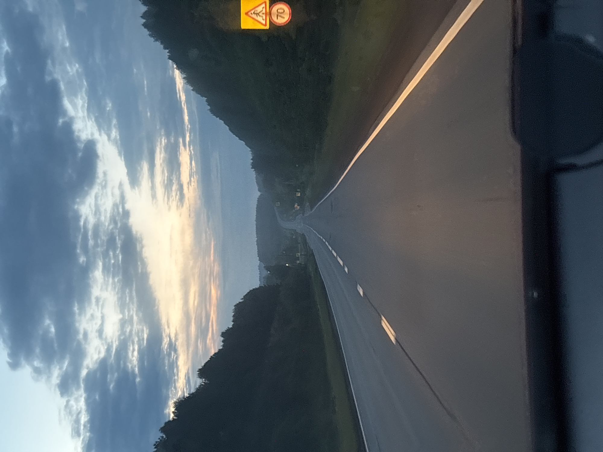 A road winds through a forested area under a sky with a combination of dark clouds and light, suggesting either dusk or dawn. A yellow road sign is visible, partially obscured by the trees, with numbers on it. The road has white lane markings and a shoulder. Part of a vehicle's interior, possibly the dashboard or pillar, is visible in the foreground, suggesting the photo was taken from inside a car. I can't verify that the location is in the city of Новая Казмаска, Russia.