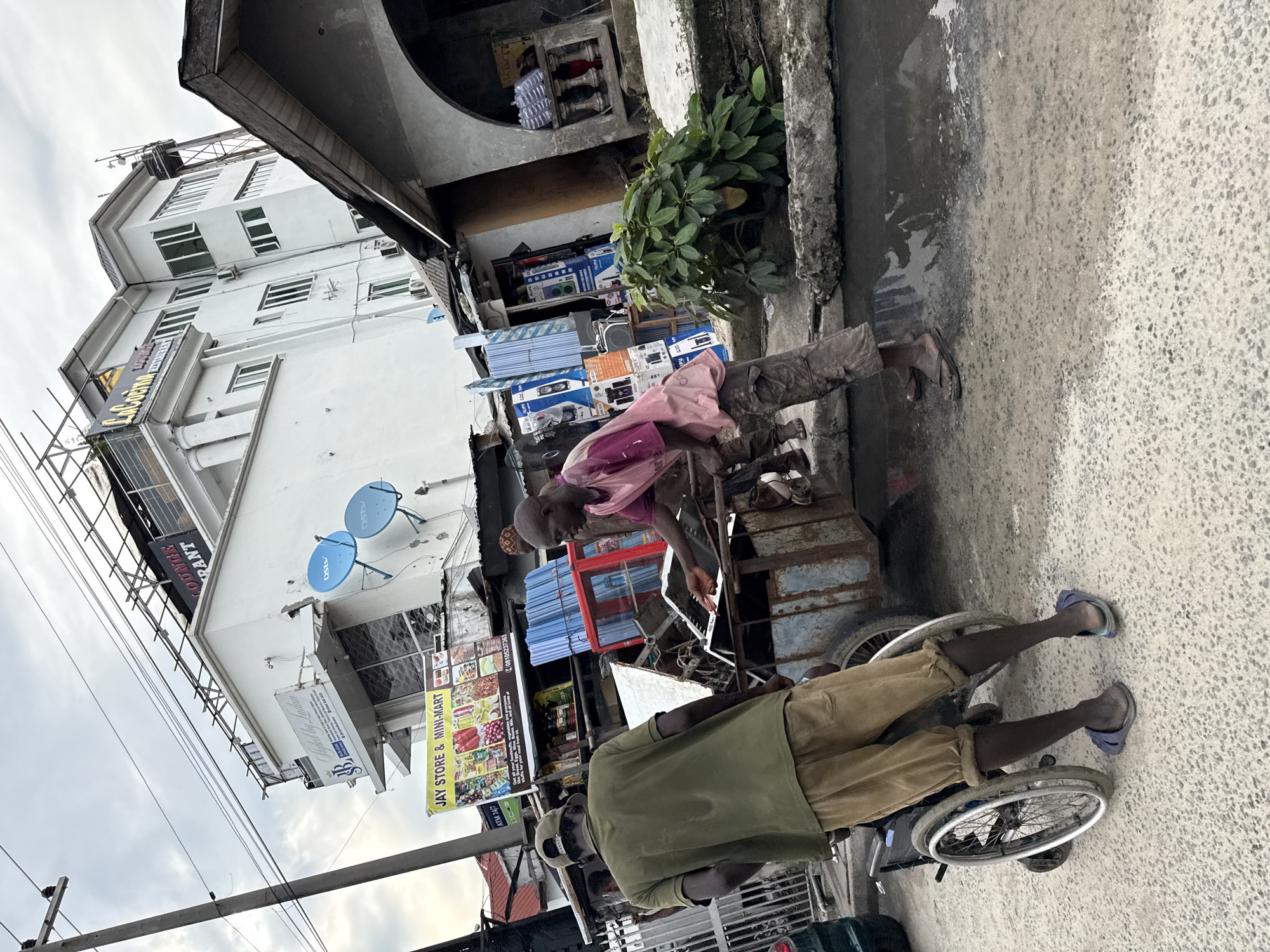 The image, rotated 90 degrees counterclockwise, depicts a street scene in Ado, Nigeria. Two men are the primary subjects. One man is in a wheelchair, wearing an olive green shirt, tan trousers, and blue sandals. The other man is pushing the wheelchair, wearing a pink shirt and dark trousers. He is pushing a cart loaded with various items, including electronics packaging and a small refrigerator.
The background features a multi-story building with white walls and multiple windows, including a sign for "Jay Store & Mini-Mart." Satellite dishes are mounted on the building's facade. Other structures include a building with a balcony with various items on it. The street is paved, with a curb visible along the edge. Power lines and poles are visible against the sky.