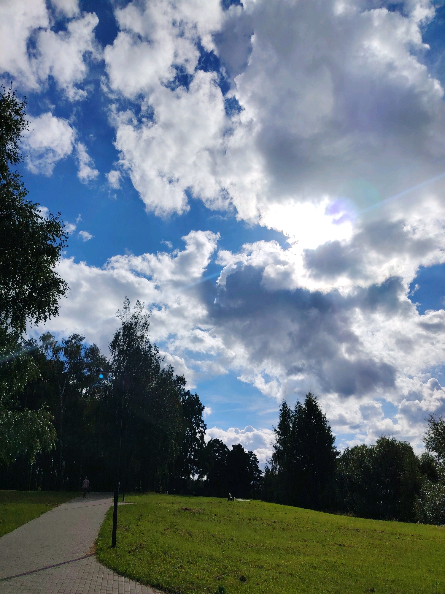 The image depicts a park scene with a paved walkway leading into a grassy area, bordered by a dense line of trees. The sky is a vibrant blue, filled with a mix of white and gray clouds. Sunlight streams through the clouds, creating a lens flare in the upper right quadrant of the image. A single person is visible walking away from the camera on the path. The overall environment suggests a peaceful outdoor recreational space, likely in Konakovo, Russia, given the context.