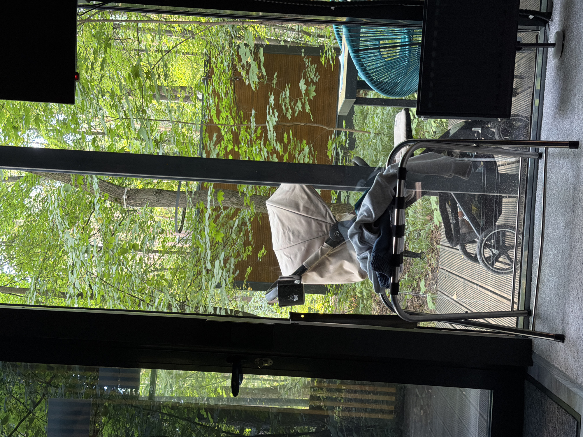 The image captures a scene through a glass-enclosed area, possibly a porch or room, with a backdrop of lush green trees. In the foreground, a metal luggage rack is set up, draped with grey and dark blue clothing. A stroller is visible behind the luggage rack, with its wheels resting on a wooden deck. To the right, a bright blue chair made of woven material stands near a table. The overall environment suggests an outdoor or semi-outdoor setting, perhaps a backyard or patio area within a wooded locale. There are no people visible in the image.