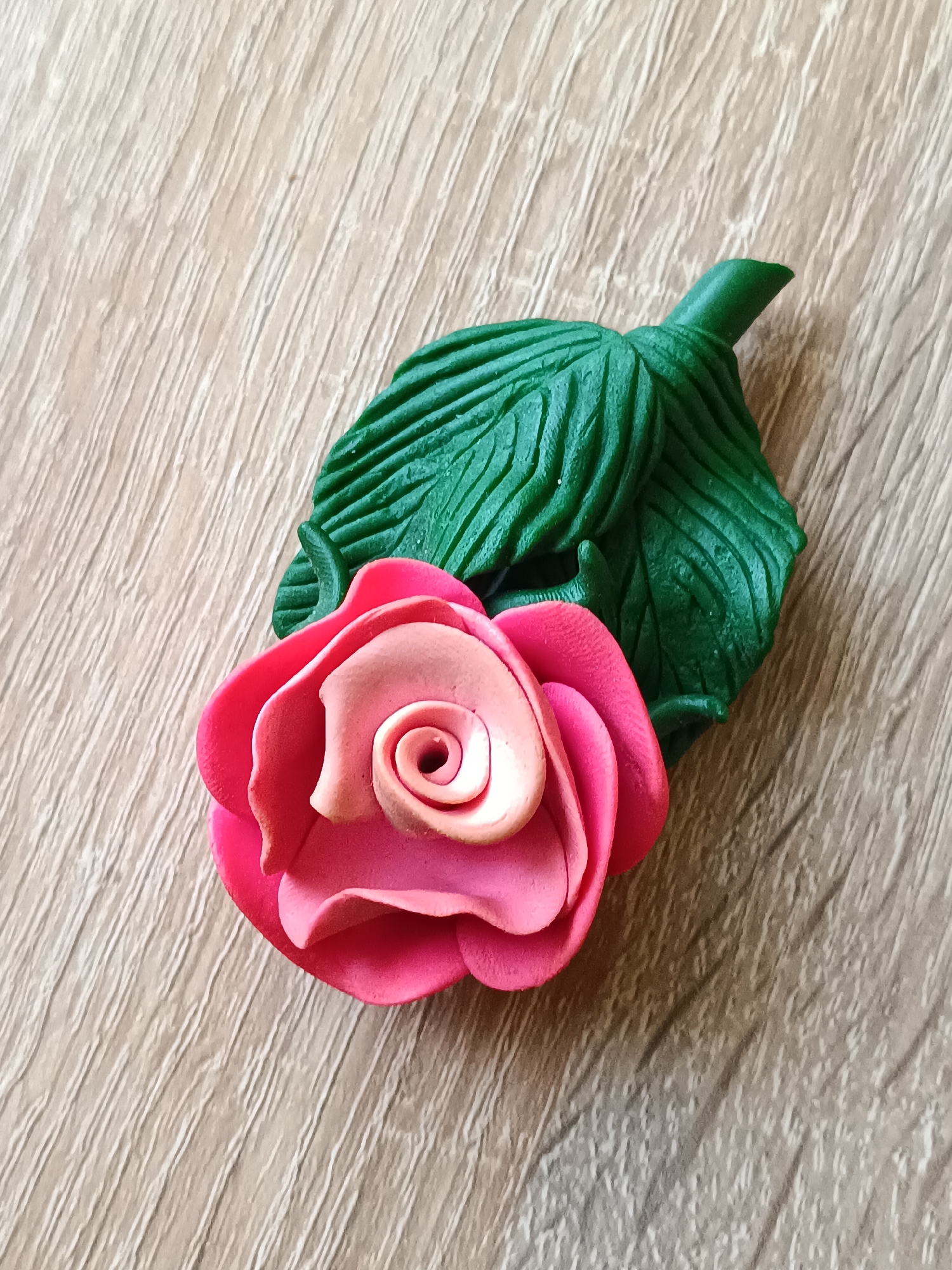 An overhead shot shows a handcrafted rose made of polymer clay, lying on a light-brown wooden surface. The rose has multiple layers of pink and light-pink petals, with a tightly wound center. Behind the rose are two green polymer clay leaves, one larger and textured with vein-like lines, and a smaller, smoother leaf emerging from the stem. The wooden surface displays a wood grain pattern with visible lines. There are no people or animals present, and the scene appears to be a still life arrangement. The location is not identifiable as being in Tebras, Latvia.