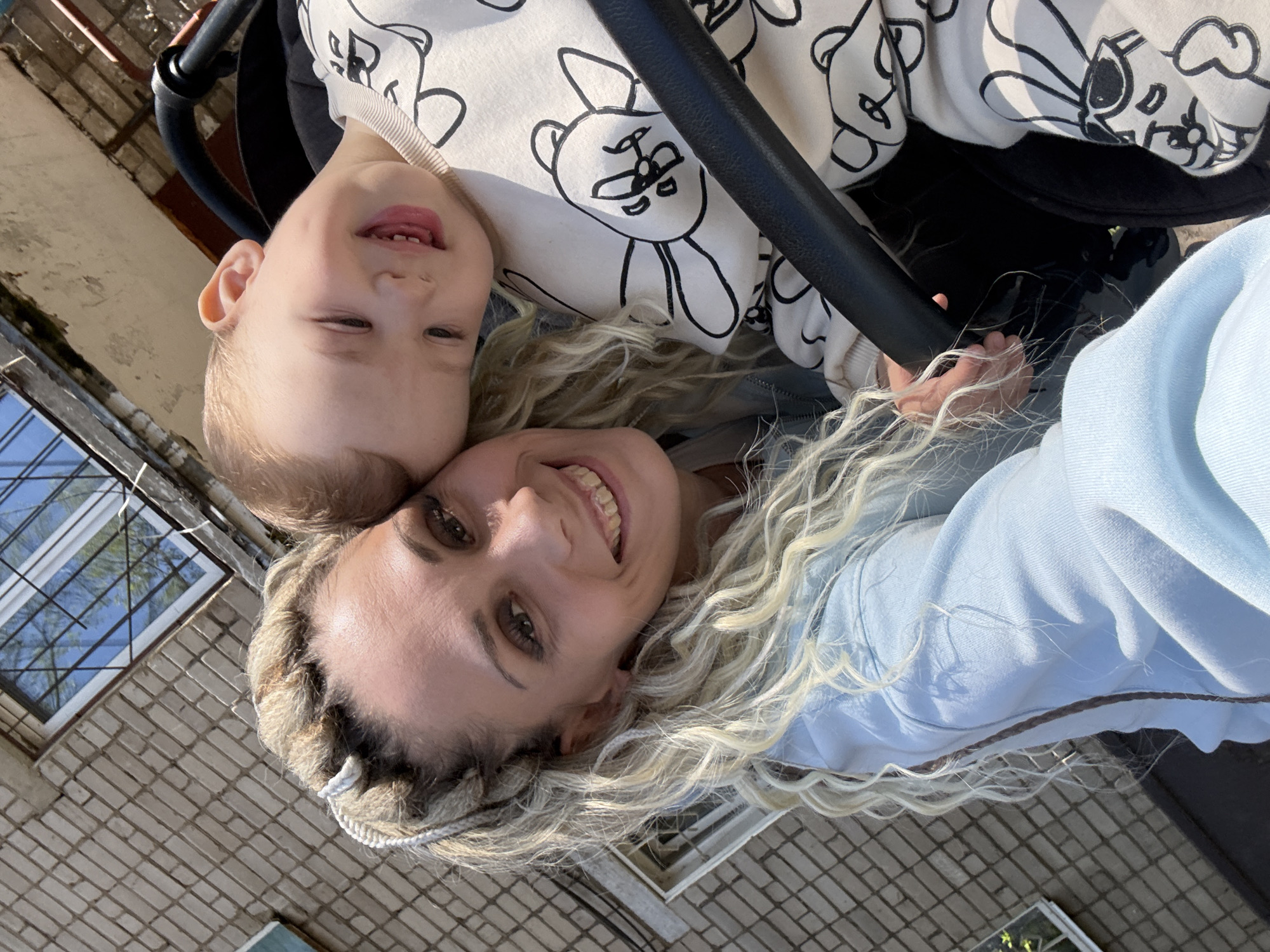 A selfie of a woman with blonde, dreadlock-style hair and a young child. The child, wearing a white top with black teddy bear prints, is in a stroller and close to the woman's face. The woman is smiling, and the child is also smiling with a missing front tooth. They appear to be outside in an urban environment, based on the brick building and barred window visible in the background. Specific location in Rusanovo, Russia, cannot be determined without further information.