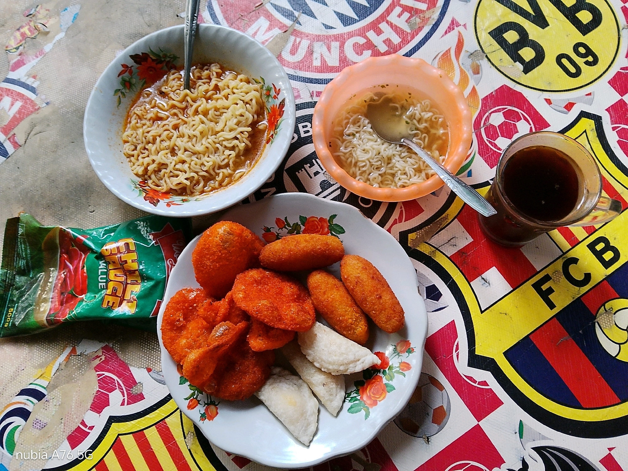 A high-angle, close-up shot captures a meal spread across a colorful, possibly laminated tablecloth featuring various sports team logos, including "BVB 09" and "FCB". The arrangement consists of a white ceramic bowl filled with instant noodles and broth, a smaller orange plastic bowl with a similar noodle dish, a white plate containing several fried, orange-colored snacks and some white, crescent-shaped items, and a clear glass filled with a dark liquid, likely coffee. A green packet of "CHILI SAUCE" is also visible. The lighting is natural and the image appears to be taken outdoors, possibly in the city of Binuang, Indonesia, given the camera's watermark "nubia A76 5G". There are no people visible in the frame.