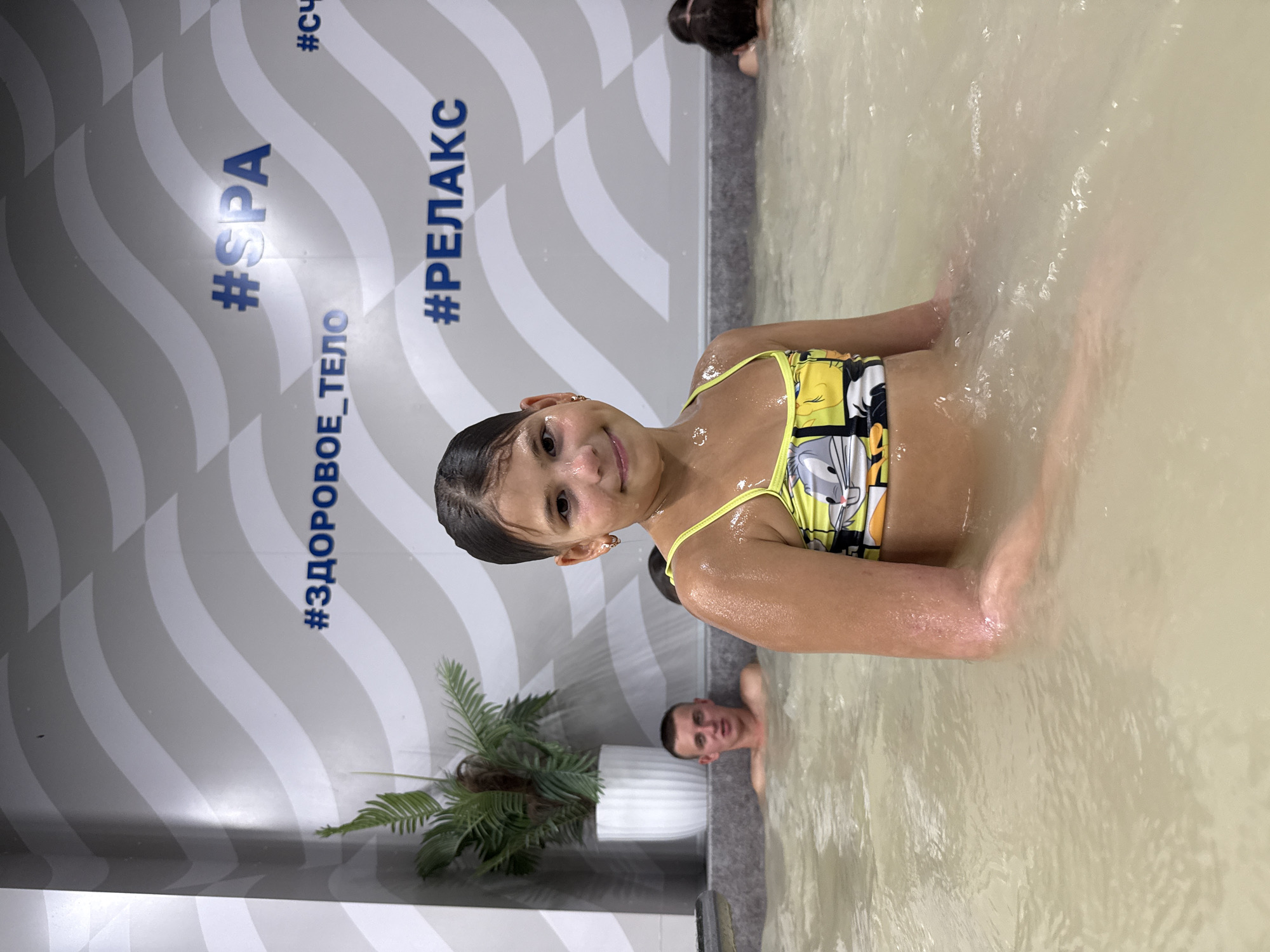 A young girl in a yellow and black swimsuit is submerged in murky water, smiling at the camera. Her hair is pulled back, and she is wearing small hoop earrings. Her arms are visible, and she appears to be enjoying herself. Behind her, a man with short hair is also in the water, looking towards the camera with a neutral expression.
The background features a wall with a wave-like pattern in shades of gray and white. Vertical text in blue is visible on the wall. On the left side of the image, a large potted plant with green fronds is placed near the edge of the water. The setting appears to be an indoor spa or pool area, possibly in Rusanovo, Russia, given the provided location context. The lighting suggests it could be daytime, with no obvious indications of weather conditions. The overall mood is relaxed and casual. The visible text on the wall includes "#SPA", "#ЗДОРОВОЕ_ТЕЛО", "#РЕЛАКС", and "#СЧ".