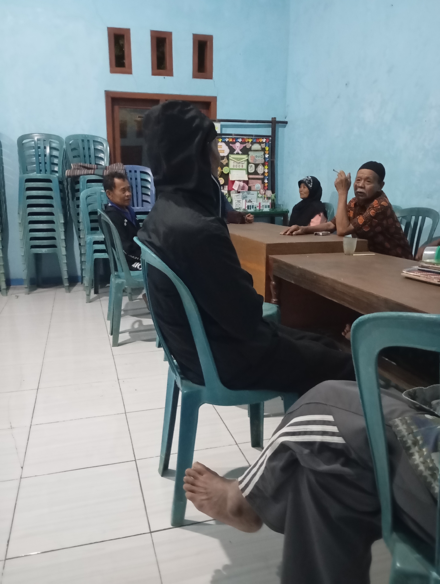 In a dimly lit room in Ketepung, Indonesia, five individuals are gathered. The room has pale blue walls and a tiled floor. Stacked blue plastic chairs are visible on the left side of the image. In the center, a person in a black hoodie sits facing away from the camera. To their right, a man in a blue shirt sits with his hands clasped. Further to the right, a woman in a black hijab and a pink shirt sits at a wooden table, and an elderly man in a patterned brown shirt and a black cap is seated next to her. He is smoking a cigarette and has his right hand raised. A glass is on the table in front of him. Another person, partially visible in the foreground, is seated on a blue chair, wearing dark pants with white stripes. Books or papers are stacked on the table in front of the elderly man. A decorative board with various items is mounted on the wall behind the group.