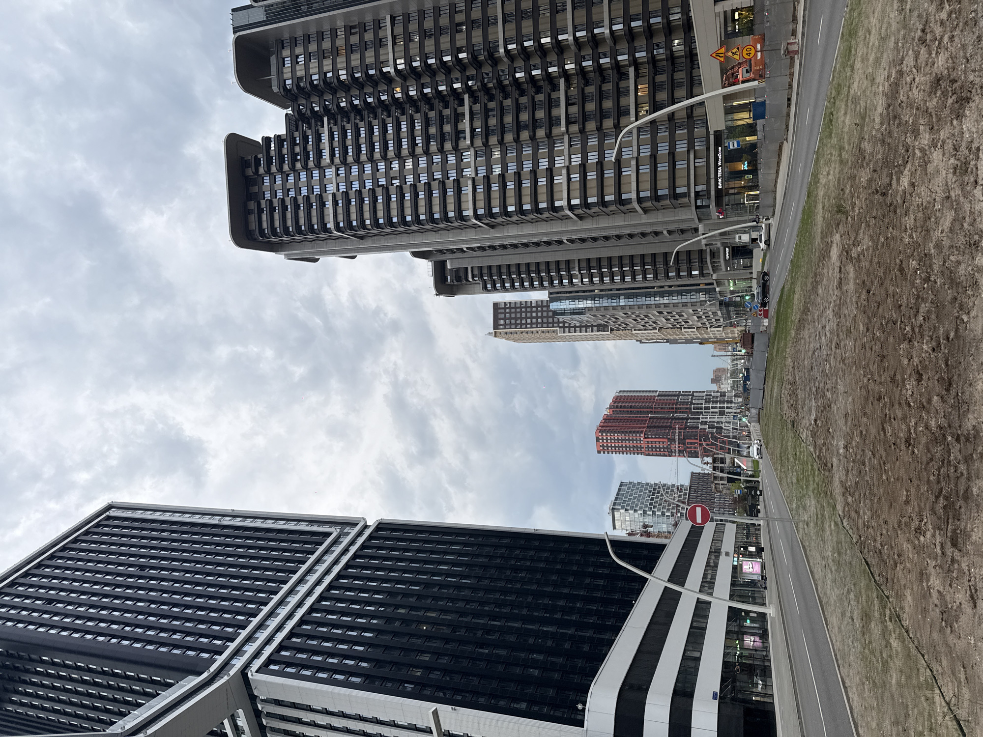 The image depicts a street scene in Moscow, Russia, characterized by modern architecture and a somewhat overcast sky. The foreground features contemporary high-rise buildings with a combination of dark-colored facades and lighter-colored structural elements. These buildings create a vertical perspective, rising towards the sky. A road and a patch of dirt or undeveloped land are visible on the right side of the image. In the background, additional high-rise buildings are visible, including one with a distinctive red and white patterned facade. The overall environment appears urban, and the composition emphasizes the architectural landscape.