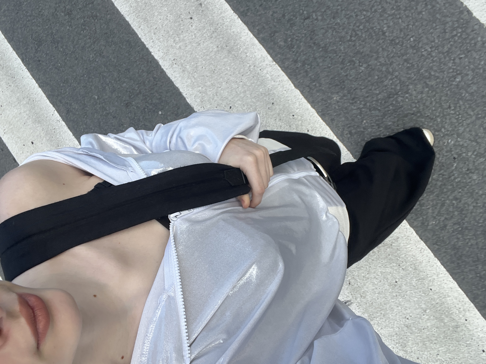 The image captures a high-angle, downward view of a person standing on a zebra crossing in Saint Petersburg, Russia. The subject's torso, one arm, and one leg/foot are visible.
The person is wearing a shimmering, iridescent white jacket or top, which is partially unzipped, revealing a black, likely strapless, garment underneath. A hand is visible holding the black strap of this undergarment. Black, loose-fitting pants cover the lower body, and a shoe with a white tip is seen on the asphalt. Part of the person's chin and mouth, adorned with light lipstick, are visible in the upper left quadrant of the frame, alongside a bare shoulder and chest area with a few small moles.
The setting is an urban street marked with a pedestrian crosswalk, featuring stark white stripes against dark grey asphalt. Bright sunlight is evident from the clear highlights on the white clothing and distinct shadows on the ground, indicating a sunny day. No specific landmarks of Saint Petersburg are visible, but the context confirms the location. There is no visible text in the image.