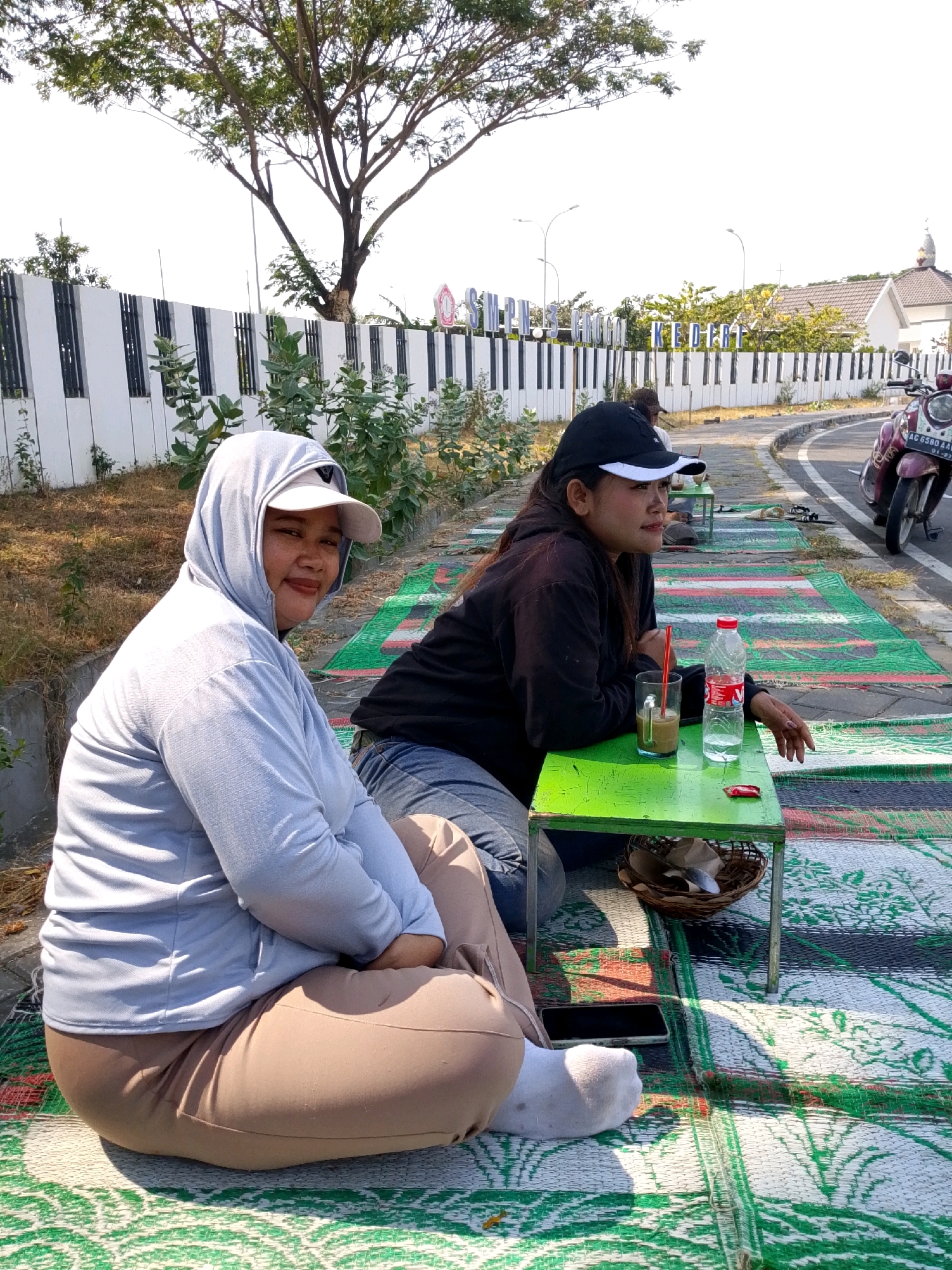 In Bulusari, Indonesia, two women are seated on mats outdoors. The woman on the left wears a light blue hooded jacket and tan pants, with a white baseball cap visible under the hood. She is looking towards the camera with a smile. The woman on the right wears a black hoodie and a black baseball cap. She is looking to her left, away from the camera. A small green table is situated between them, holding a glass of coffee with a red straw, a bottle of water, and a small red candy. A motorcycle is parked on the right side of the frame. In the background, a white fence with black vertical bars runs along a paved road, and trees and buildings are visible. The ground is covered with a colorful mat.