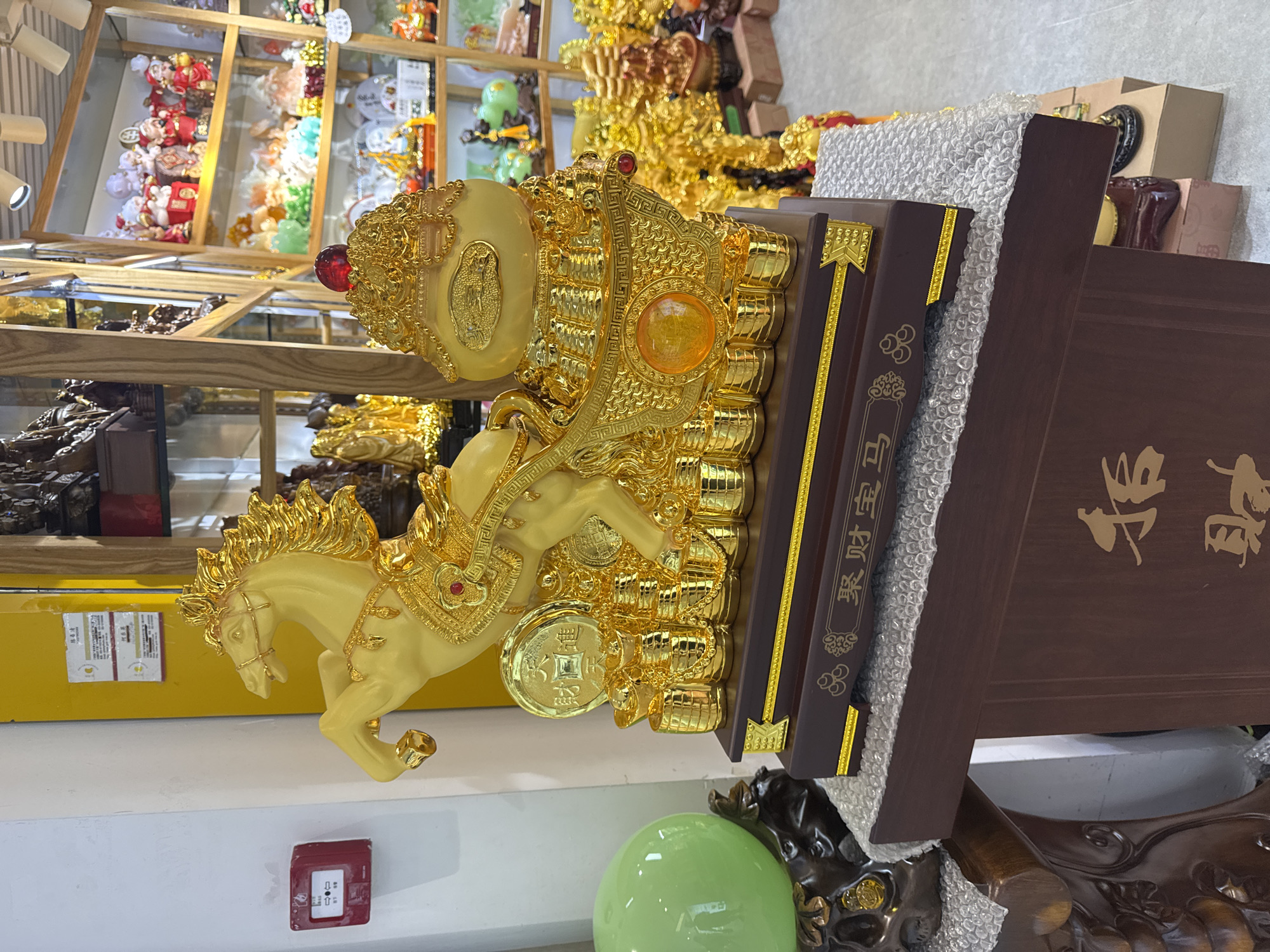 The image shows a golden horse statue displayed indoors. The statue is mounted on a dark brown wooden base with Chinese characters inscribed on it. The backdrop includes shelves stocked with various decorative items, suggesting the statue is located in a shop or store. The presence of what appears to be a fire alarm pull station on the wall further indicates an indoor commercial setting. The presence of Chinese script on the wooden base and metadata (likely indicating the picture was taken) suggests the location is Yuexiu District, China.