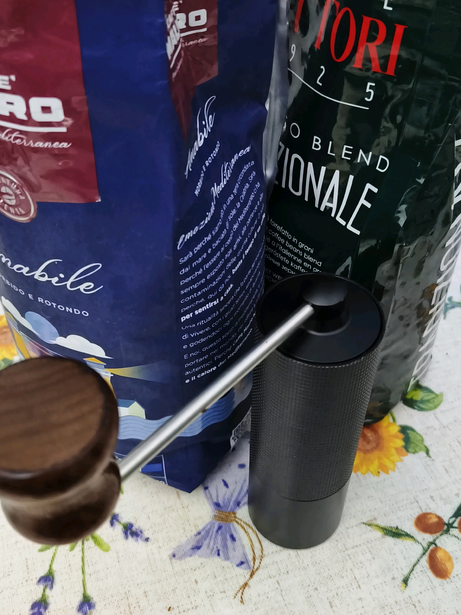 The image shows two bags of coffee and a manual coffee grinder on a patterned tablecloth. One bag is dark green with red and white lettering that reads "TORI 9 2 5 O BLEND ZIONALE". The other bag is dark blue with white lettering that reads "RO Mediterranea" and other text in Italian. The coffee grinder is black with a silver handle and a wooden knob. The tablecloth has a floral pattern with sunflowers and lavender. The scene is an indoor setting, likely a kitchen or dining area. There are no people visible in the image. There is no indication that this scene is located in Livorno, Italy.