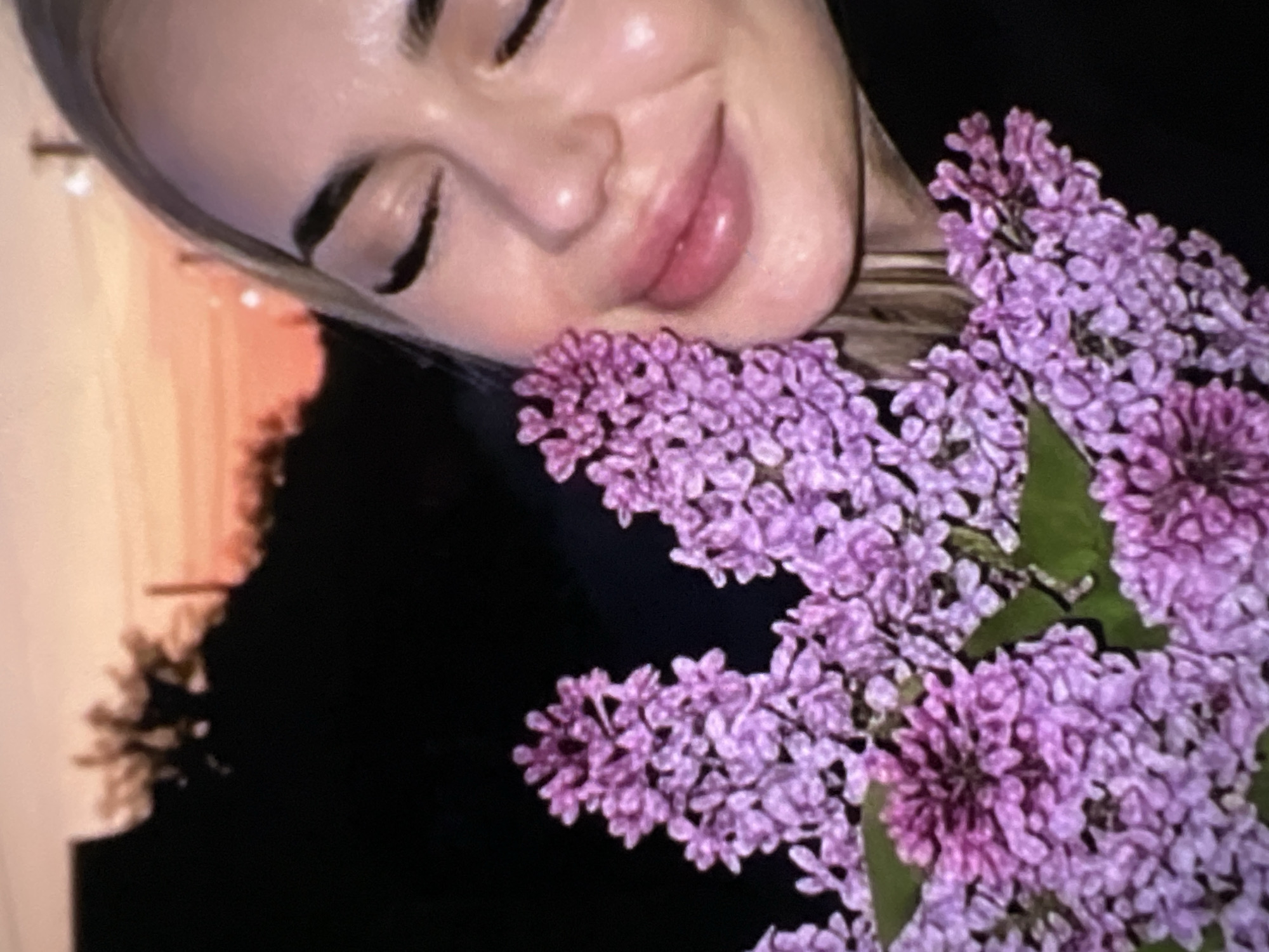 The image captures a close-up, rotated view of a person with a large bouquet of lilac flowers. The subject is a woman, seen from the nose down to the chin, with her eyes closed and a gentle smile on her lips. Her skin appears smooth and well-lit, and her lips are full with a glossy, light pink hue. Dark eyebrows and possibly eyeliner/mascara are visible on her closed eyelids. Her hair, light in color, frames the left side of her face.
A dense cluster of vibrant purple and lavender lilac blossoms occupies the foreground and extends across the right side of the frame, with some green leaves interspersed. The flowers appear fresh and are held close to the woman's face, suggesting she is enjoying their scent or beauty.
The background on the far left shows a soft, warm orange glow, indicative of either a sunrise or sunset, with silhouetted tree branches or foliage against it. The rest of the background is predominantly dark, creating a strong contrast that highlights the illuminated face and the colorful flowers. No specific location cues are visible. The overall mood conveyed is one of tranquility, happiness, and appreciation for nature. There is no visible text in the image.
