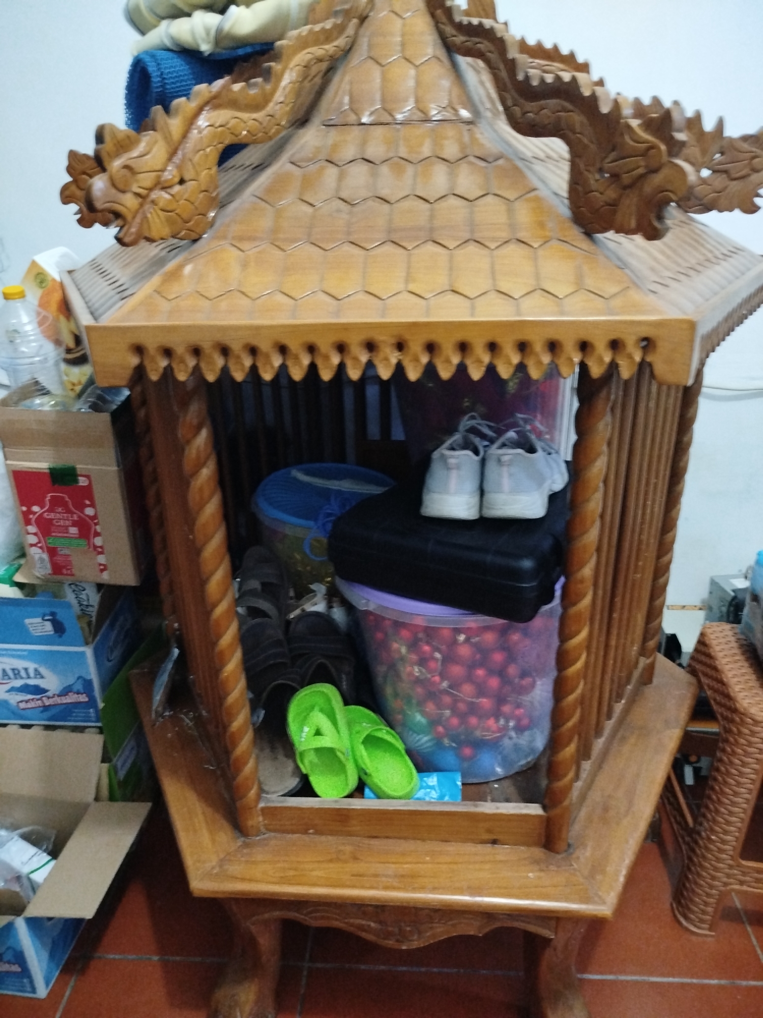 A medium shot captures a wooden structure resembling a shrine or decorative display cabinet, intricately carved with dragon motifs on its roof. The structure is positioned indoors, with a tiled floor visible in the foreground and a plain wall in the background. The cabinet contains various objects, including a pair of light grey sneakers placed on a black case, a blue bucket, and a transparent plastic container filled with red spherical objects, possibly ornaments or fruits. Several pairs of sandals are also present. To the left of the cabinet, cardboard boxes are stacked, one of which displays text and imagery suggesting a product packaging. The lighting suggests an indoor environment with ambient light. The scene appears to be a storage area or a display within a home or shop.