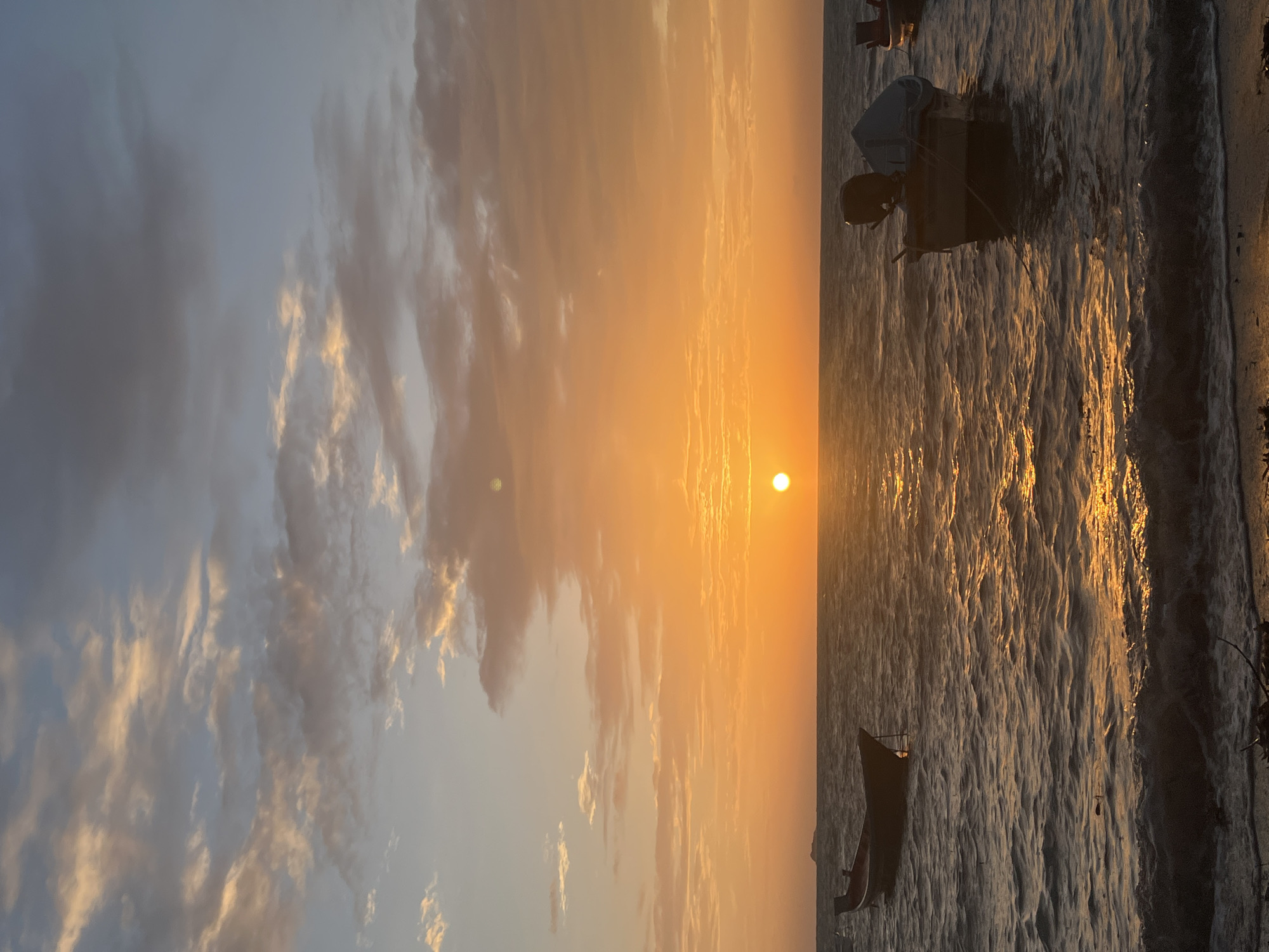 This is a vertical, full shot of a sunset over the ocean, likely taken in the Ko Pha-ngan District of Thailand. The sun, a bright orange orb, sits just above the horizon, casting a golden, shimmering reflection on the choppy water. The sky is a gradient of orange, yellow, and pale blue, with scattered clouds tinged with pink and grey.
In the foreground, the dark sand of a beach is visible, with some scattered debris. Several small, dark boats are moored in the water. One boat is on the left, closer to the shore, and appears to be empty. Another boat is to the right, with a dark outboard motor and a grey cover. The water is a deep blue, with white foam and ripples near the shore. The overall impression is one of serene natural beauty as the day ends.