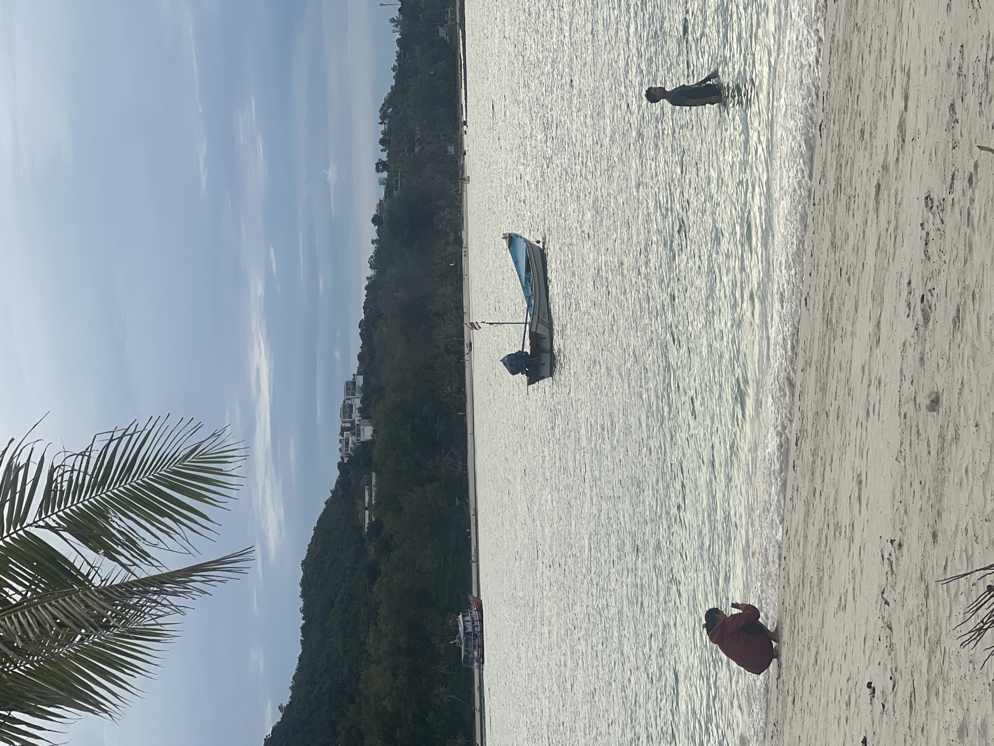 The image depicts a serene beach scene in the Ko Pha-ngan District of Thailand. In the foreground, a sandy beach stretches across the bottom of the frame, with a few scattered pieces of driftwood. At the edge of the water, a person wearing a dark red jacket is crouched down, appearing to be taking a photograph with a mobile phone. To the right, a person stands in the shallow water, facing away from the camera.
The sea occupies the middle ground, with gentle waves lapping the shore. A small, light-colored boat with a blue trim is anchored in the water, and a Thai flag is visible on its mast. Further out, a larger red and white boat is visible near the shore.
In the background, a lush green, tree-covered hillside rises steeply. Several modern white buildings are nestled on the slope of the hill. The sky above is overcast with a soft, diffused light. A palm frond hangs down from the upper left corner of the frame, adding a tropical element to the scene. The overall atmosphere is tranquil and peaceful, suggesting a quiet day at the beach.