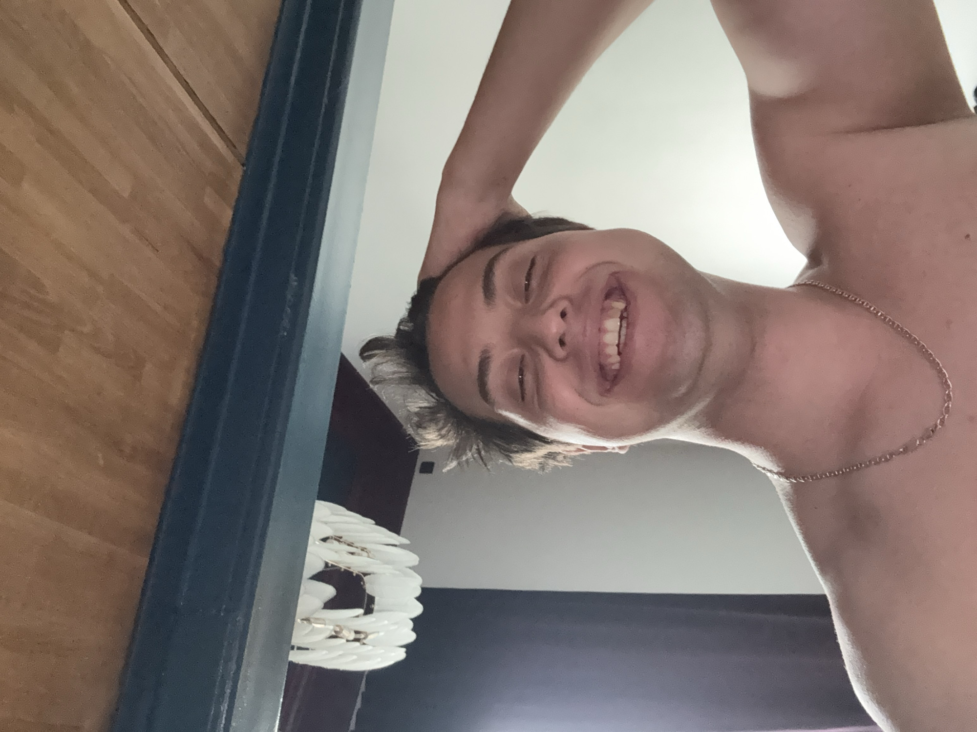 This image captures a young man in an indoor setting, likely a home in Vidnoye, Russia. The photograph is taken from a low-angle perspective, looking up at the subject. The man is shown from the shoulders up, smiling broadly with his teeth visible, conveying an emotion of happiness or cheerfulness. His left arm is raised, with his hand placed on the back of his head. He appears to be shirtless, and a thin gold chain is worn around his neck.
To the left of the frame, a wooden surface, possibly a floor or part of a wall, is visible, bordered by a dark blue vertical doorframe or trim. In the lower-left background, a distinctive white, modern light fixture with multiple petal-like or feather-like elements is partially seen. The rest of the background consists of a light-colored ceiling and a darker wall section. No visible text is present in the image.