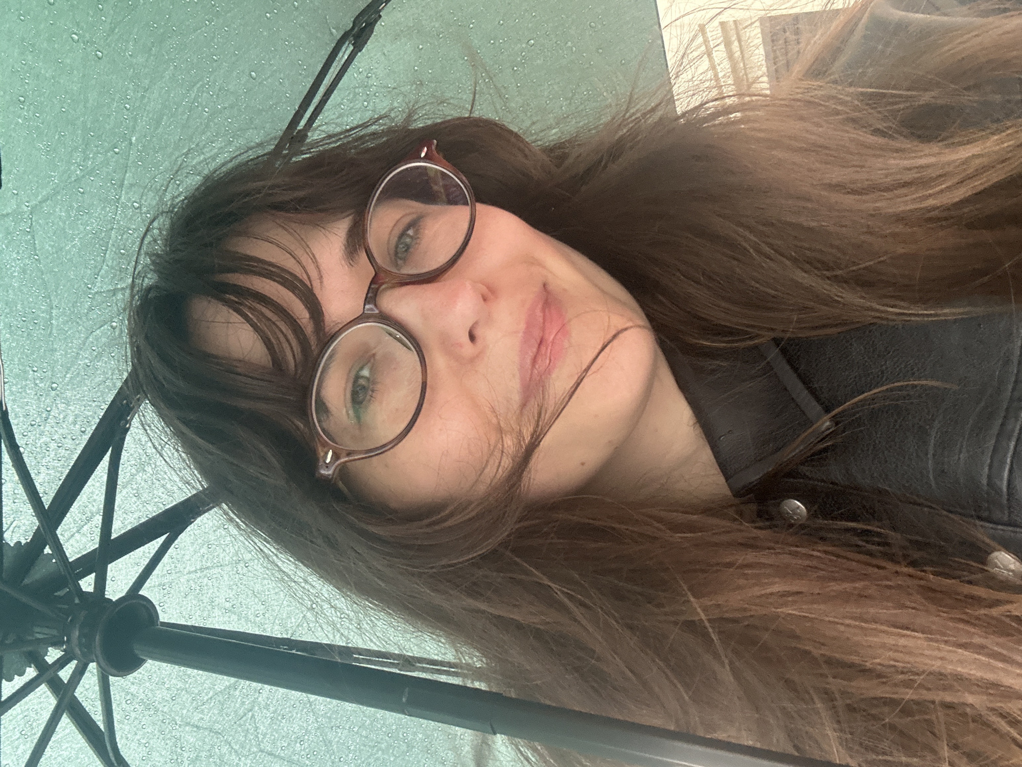 A selfie taken from below a teal umbrella reveals a woman with long, brown hair and round, brown-rimmed glasses. She is wearing a black leather jacket with visible buttons. The umbrella's fabric is wet, with raindrops visible. The background is out of focus and appears to be a street or building facade. The lighting suggests an overcast or rainy day. The image quality is average, with visible noise.