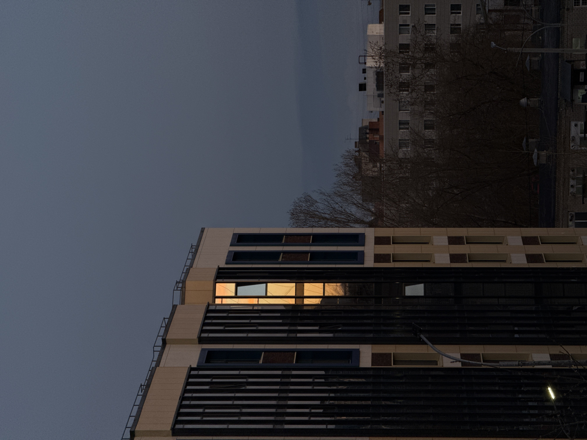 The image shows a nighttime urban scene. A modern building with a façade of beige panels and dark, horizontal brise-soleils occupies the foreground. The windows reflect a warm, orange light. In the background, there is a typical Soviet-era apartment building. The sky is a dark, overcast gray. Bare tree branches are visible. This is likely a scene in the city of Perm, Russia, judging by the architecture and general urban character.