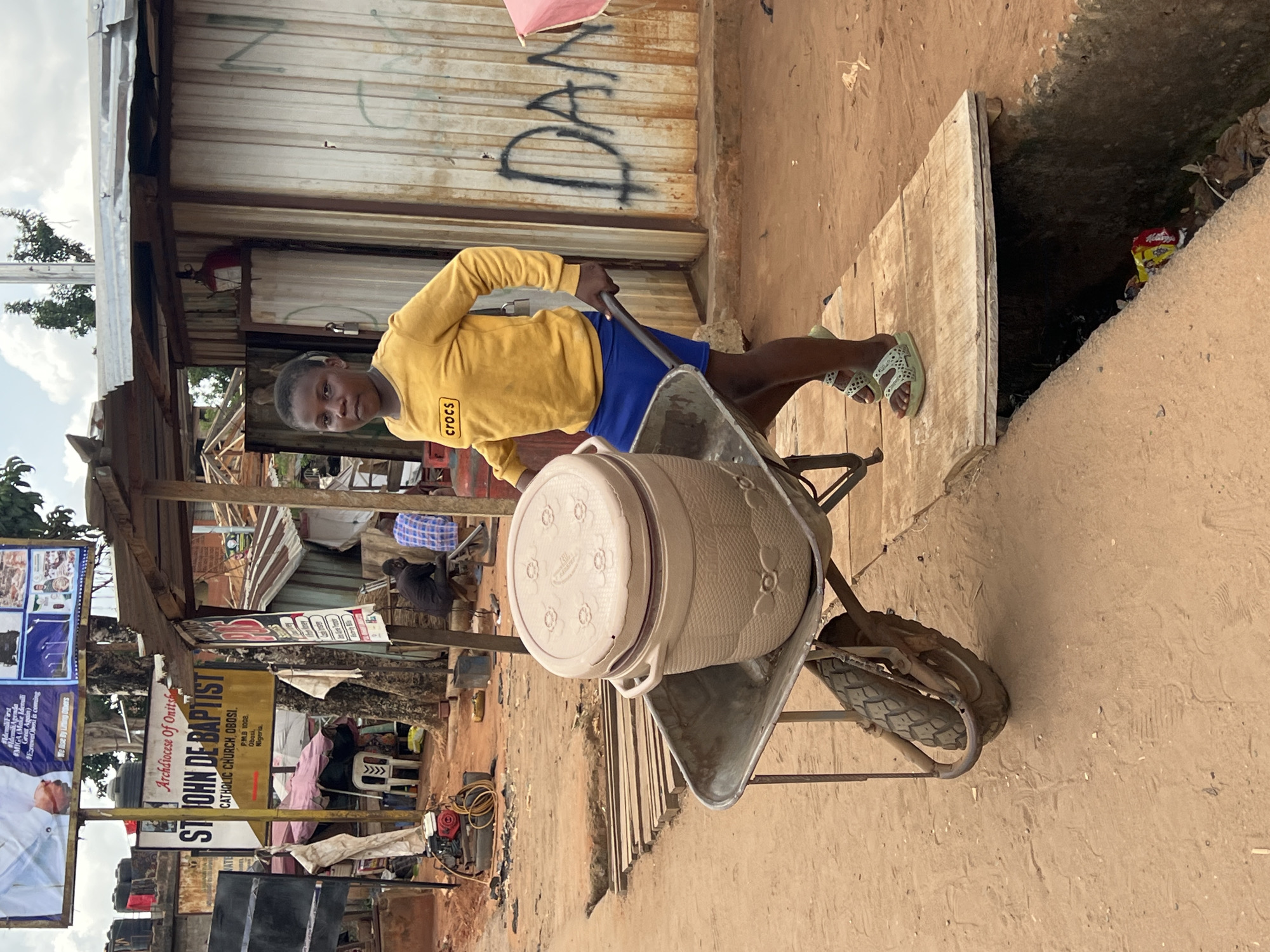 A young man wearing a yellow Crocs sweatshirt and blue shorts stands next to a wheelbarrow holding a large, covered container. He is looking at the camera. The wheelbarrow is positioned next to a dark opening in the ground, possibly a sewer or drainage. Behind the man, a metal building with graffiti reading "DAN" is visible. In the background, faded signage for St. John De Baptist Catholic Church, Archdiocese of Onitsha, in Obosi, Nigeria, is visible. The overall environment appears to be a street or roadside in Onitsha, Nigeria, possibly a slum, with a mix of temporary structures and signs.