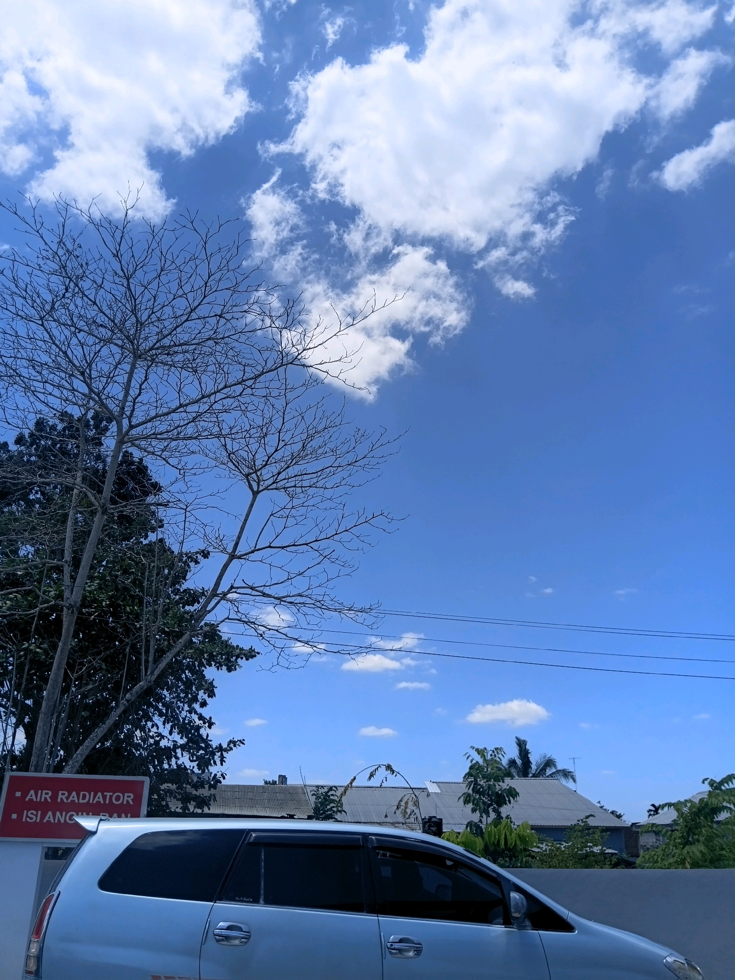 A full-shot vertical photograph captures a bright, sunny day with a clear blue sky and scattered white clouds. In the foreground, the rear section of a light-colored minivan is visible, with its tinted windows and door handle in view. To the left of the minivan, a portion of a red sign with white text is partially visible. Above the sign, the branches of a bare tree, devoid of leaves, extend upwards into the sky. A densely leaved, dark green tree stands to the left of the bare tree. Further back, a series of rooftops with metal coverings are visible, suggesting buildings in a suburban or semi-urban area. Electrical wires are strung across the upper part of the frame. The scene appears to be set outdoors, possibly on a street or in a residential area in Sungai Kapitan, Indonesia, given the presence of the specific sign. There are no people or animals visible in the frame.
