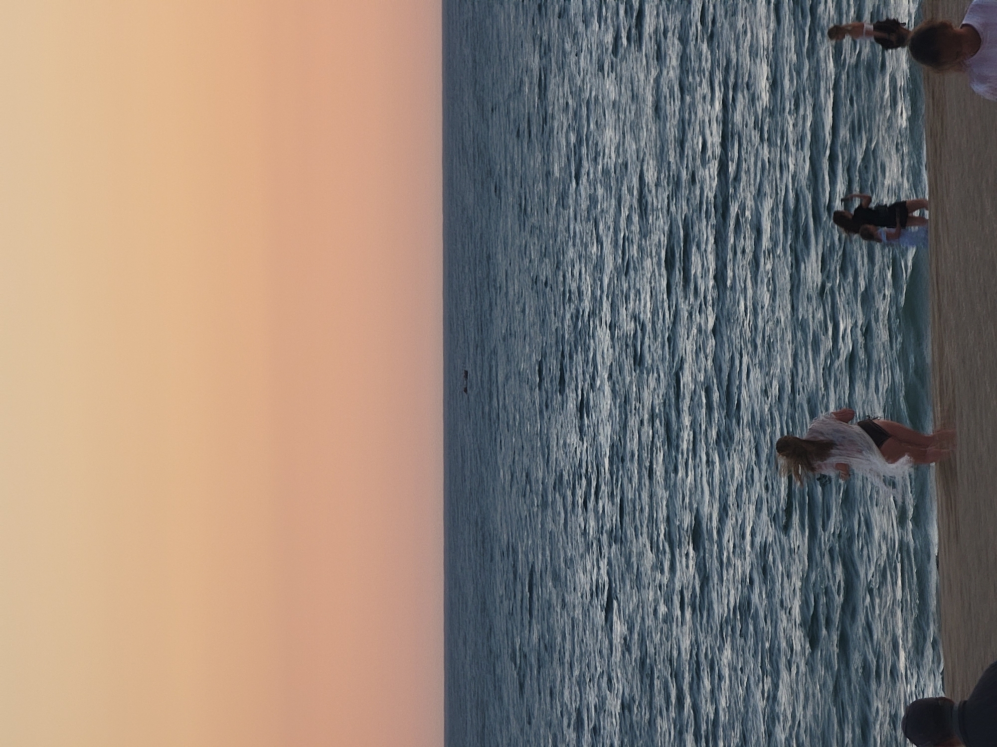 An aerial perspective captures a serene beach scene at dusk in Quarteira, Portugal. The sky is a soft gradient of peach and orange, transitioning into the deep blue of the Atlantic Ocean. Waves gently lap against the sandy shore where several individuals are present. In the foreground, a person's back is visible, facing the water. Further down the beach, a woman in a white cover-up and black bikini runs towards the sea. To her right, two people stand near the water's edge, one of whom appears to be taking a photograph. On the far right, another person is visible. In the distance, a small boat is silhouetted against the horizon.