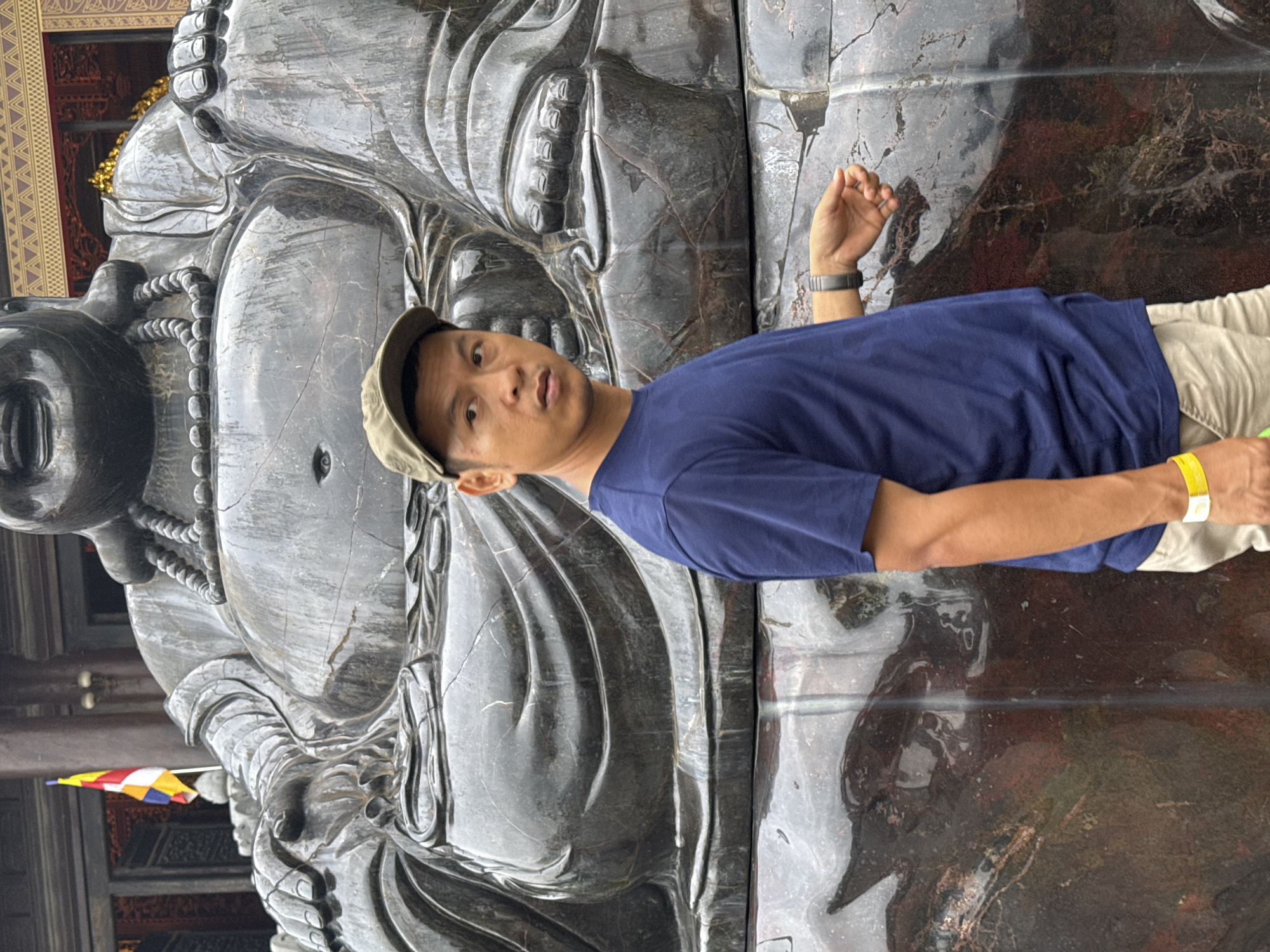 A man wearing a blue t-shirt and a green baseball cap stands in front of a large stone statue of Buddha. The statue is dark grey and appears to be made of polished stone, with intricate details carved into the fabric of the Buddha's robe and a beaded necklace around its neck. The man is looking off to the left, with his right hand extended forward, fingers splayed. He is wearing a watch on his left wrist and a yellow wristband on his right. In the background, a multi-colored flag can be seen attached to a pole. The ground in front of the statue appears to be made of stone as well, with a glossy, wet appearance in some areas. The scene is likely located in Tây Hoa Lư Ward, Vietnam, given the presence of the Buddha statue and the flag, which resembles the Buddhist flag.