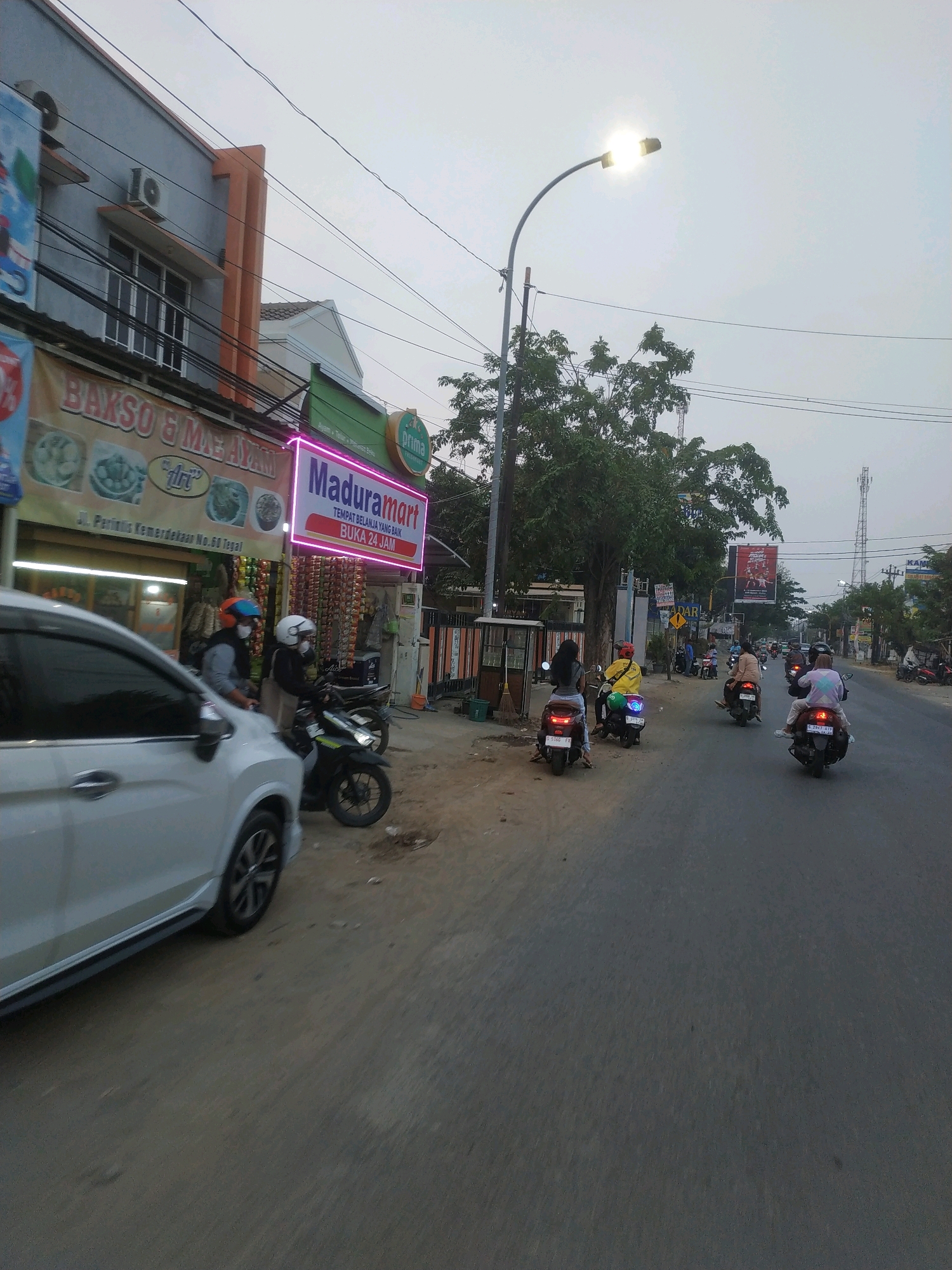 A street scene in Tegal, Indonesia, captures a moderate amount of traffic. A white car is prominently displayed on the left side of the frame, its front portion blurred due to motion. Several motorcycles are also present, some carrying single riders and others with passengers. The motorcycles are traveling in the same direction as the car. On the right side of the street, there are buildings, including a store with a prominent neon sign that reads "Maduramart" and displays text in Indonesian. Other shops and businesses are visible in the background, with some appearing to sell goods like food and beverages. A streetlamp illuminates the scene, and utility wires are strung across the sky. The overall atmosphere suggests a typical day in an urban environment with ongoing commercial activity and vehicular movement.
