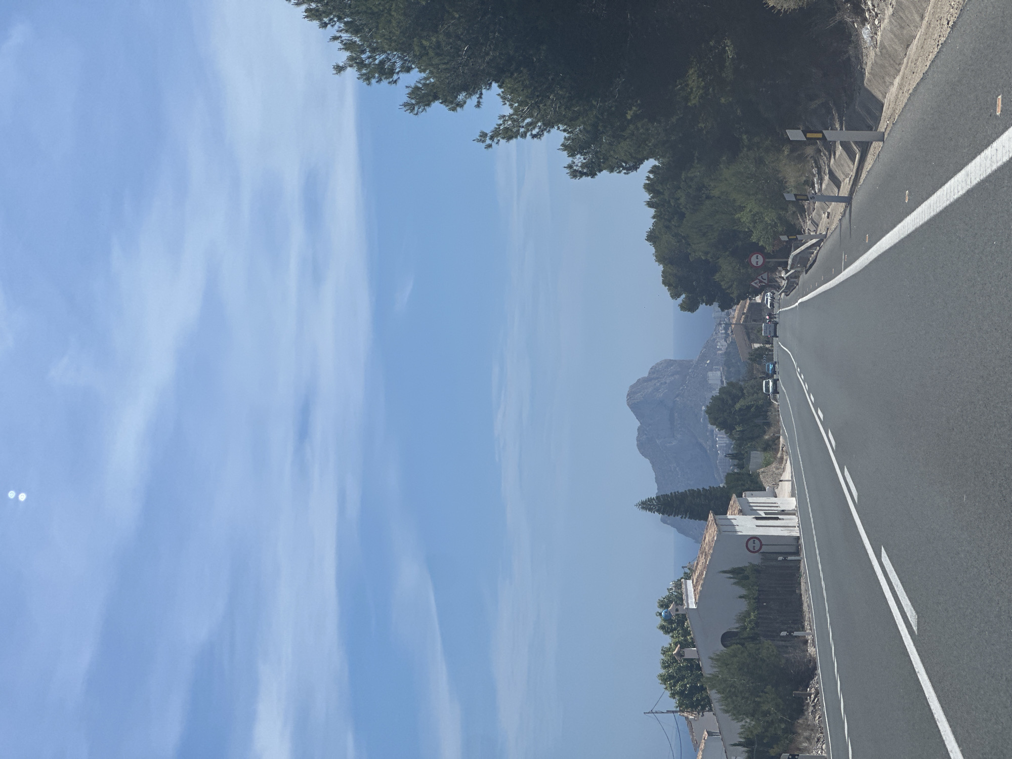 The image shows a road in Benissa, Spain, with the Peñón de Ifach visible in the background. The road has white lane markings, and there are several vehicles on it. Trees line the side of the road. Buildings are seen along the roadside. The sky is blue with some clouds.