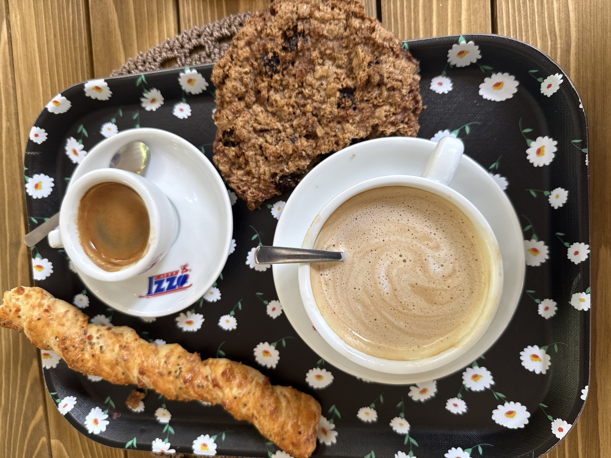 A black tray with a floral pattern of white daisies with red centers and green leaves sits on a wooden table. On the tray are two cups of coffee, one espresso and one cappuccino, a twisted breadstick with seeds, and a large cookie. The espresso cup is white with a red and blue logo that reads "OZZI". A small spoon rests on the saucer of the espresso cup. The cappuccino has a foamy top and a spoon rests in it. The breadstick is golden brown and appears to be made of puff pastry. The cookie is large, round, and has a textured surface with visible raisins or dried fruit. The scene appears to be set at a cafe in Stara Zagora, Bulgaria, based on the "OZZI" logo on the espresso cup, which is a known cafe chain in Bulgaria.