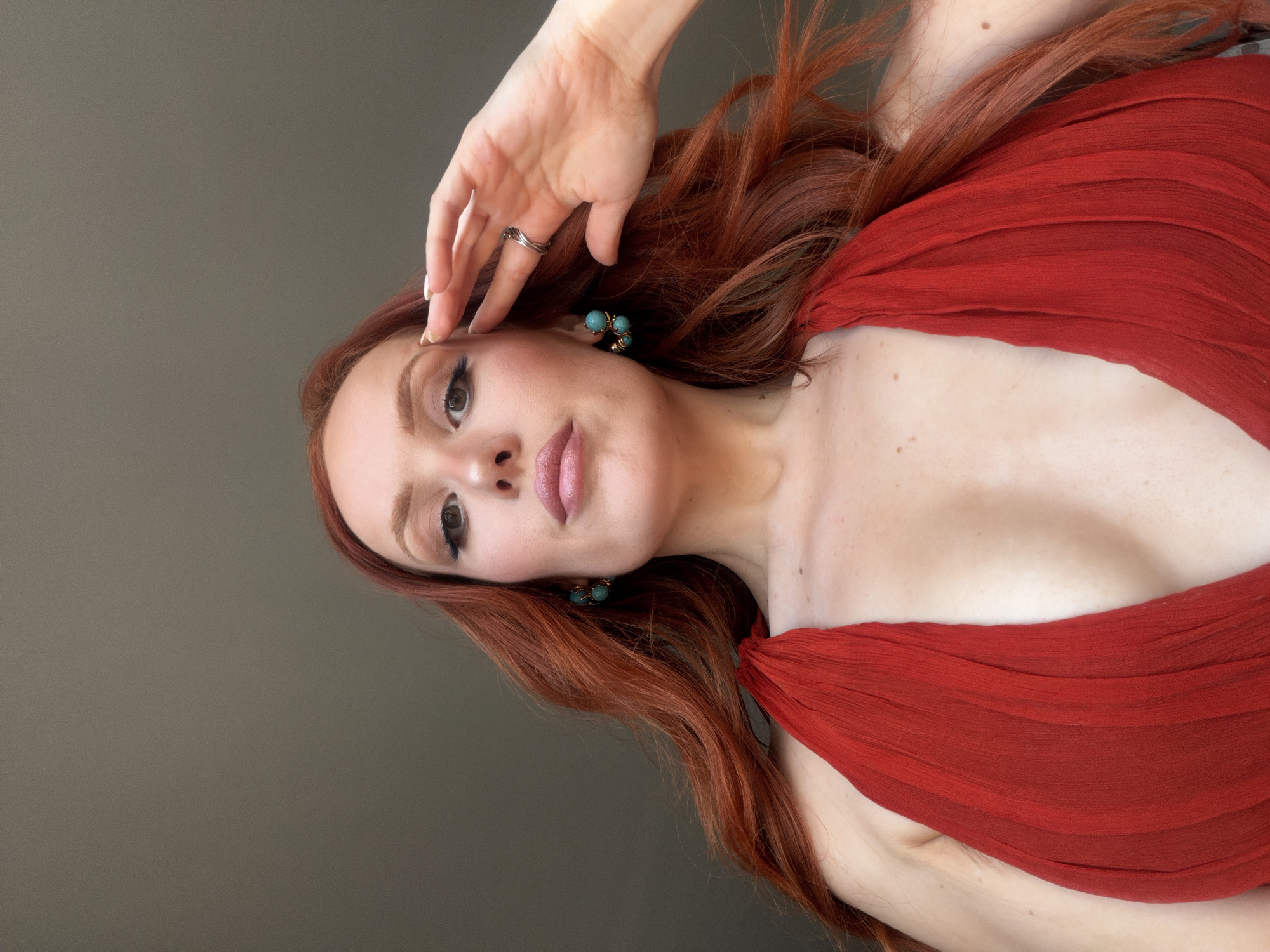 A woman with long, red hair is lying on her side, facing the camera. She is wearing a red dress or top with a draped neckline. Her left hand is near her forehead, with a silver ring on one of her fingers. She has light skin, brown eyes, and is wearing makeup, including lipstick. She is wearing earrings with turquoise-colored stones. The background is a solid gray-green color. The location is not identifiable as being in Paris, France.