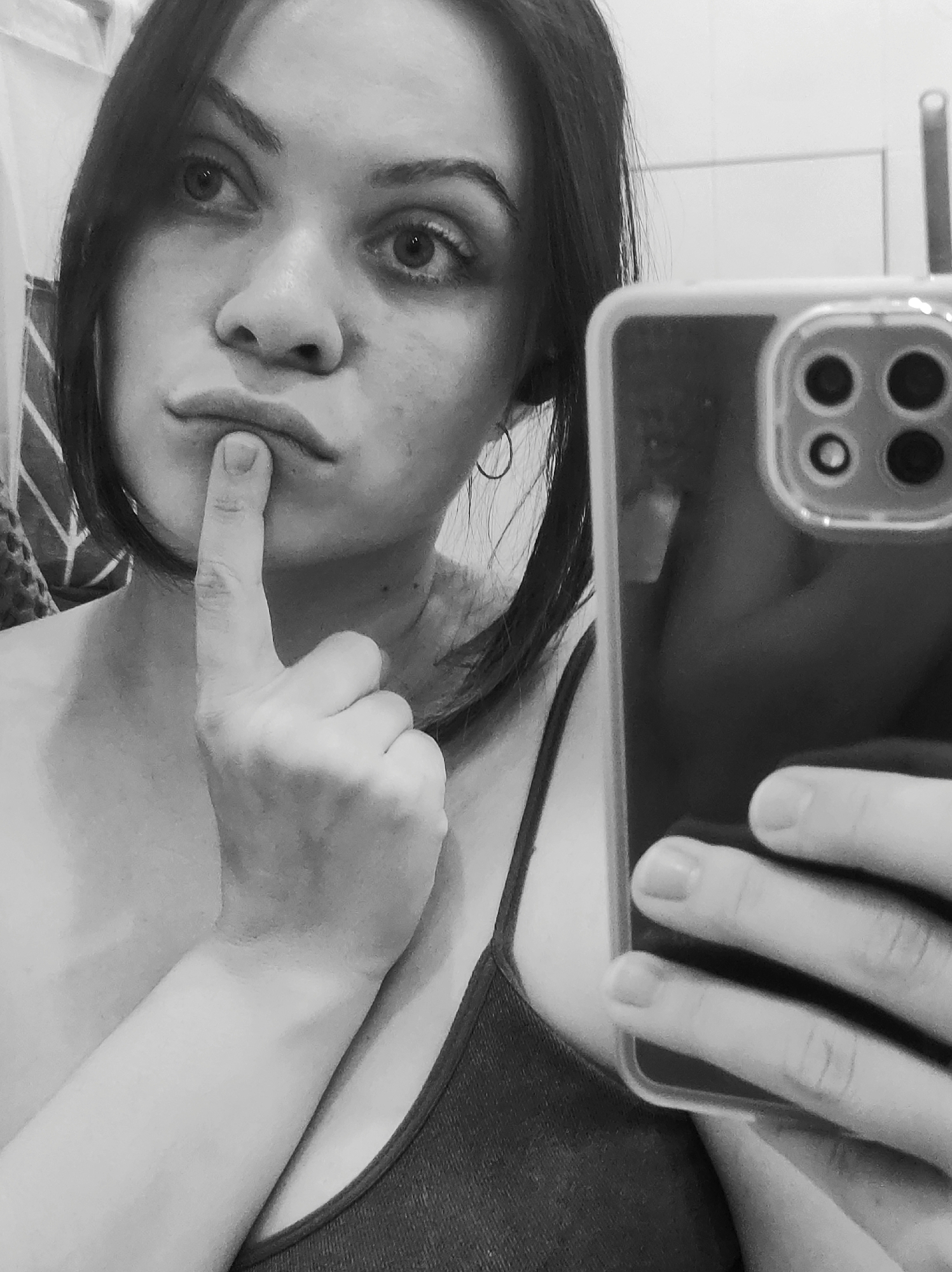 A monochrome mirror selfie depicts a young woman, captured from the chest up. She presents a thoughtful or questioning expression, with her right index finger placed against her slightly pursed lips. Her wide, dark eyes are cast upwards and to the right, conveying a sense of contemplation, surprise, or a playful "shhh" gesture. Her dark hair frames her face and falls around her shoulders. She wears a dark-colored spaghetti-strap top, and a simple hoop earring adorns her left ear.
Her left hand holds a smartphone, partially visible on the right side of the frame, indicating she is taking the photo in a mirror. The phone is enclosed in a clear case, and its prominent multi-lens camera module is clearly visible. The background, reflected behind her, is a light-toned, indistinct indoor setting, possibly a tiled wall of a room or bathroom, common in residences in locations like Chaikovsky, Russia. The black and white aesthetic emphasizes the emotional nuances of her facial expression and the textures within the image. No visible text is present. The time of day and weather are not discernible.
