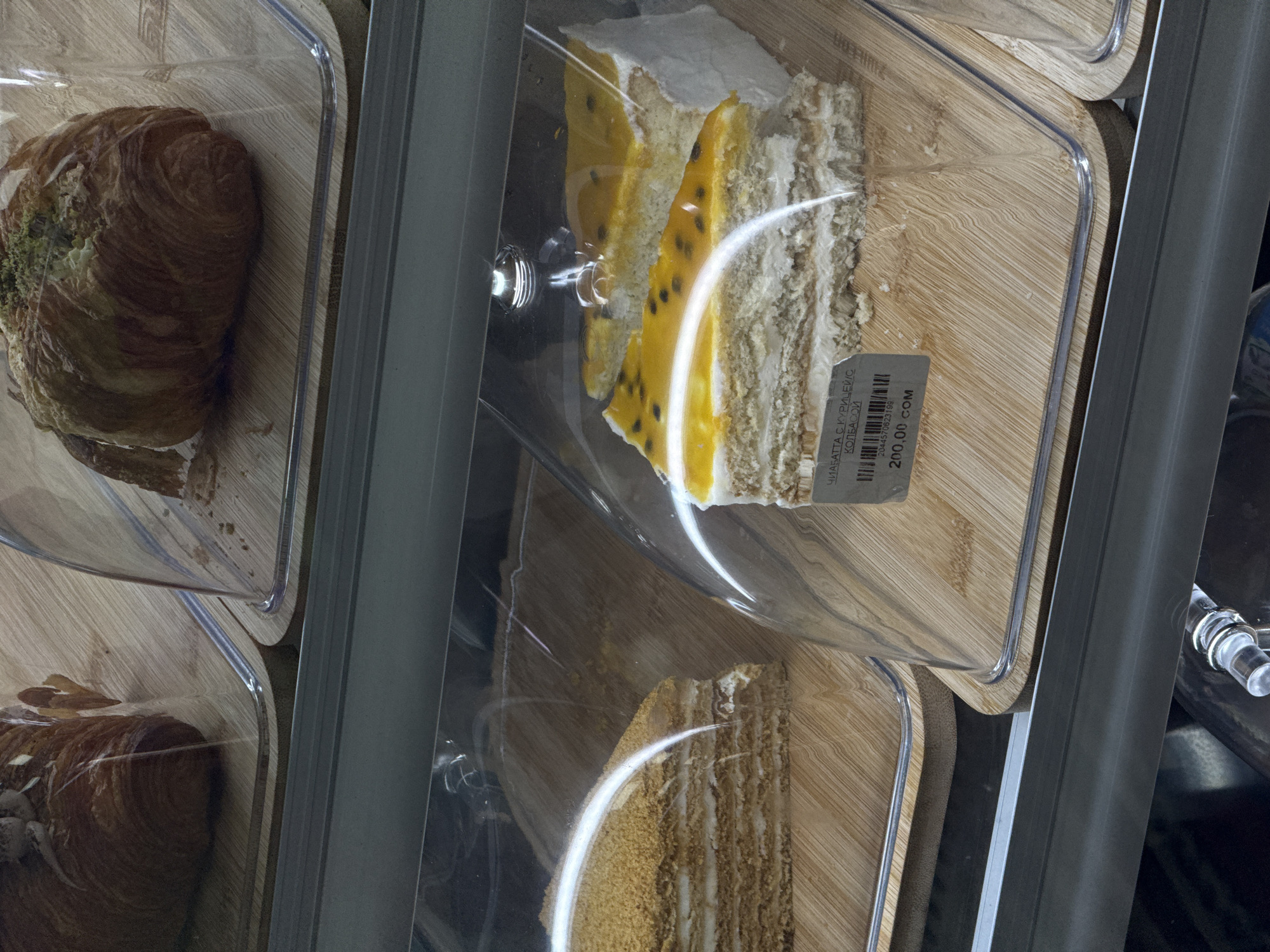 An overhead shot shows two croissants and two slices of cake displayed in a refrigerated case at a bakery in an unknown location. The croissants are on wooden boards covered with plastic domes. One croissant is golden brown and flaky, with some green specks on top. The other croissant is also golden brown. Two slices of cake are on a wooden board also covered by a plastic dome. The cake slices are layered with cream and topped with a yellow glaze with black seeds. A label on the cake board reads "200,00 сом". The refrigerated case appears to be made of metal and glass.
