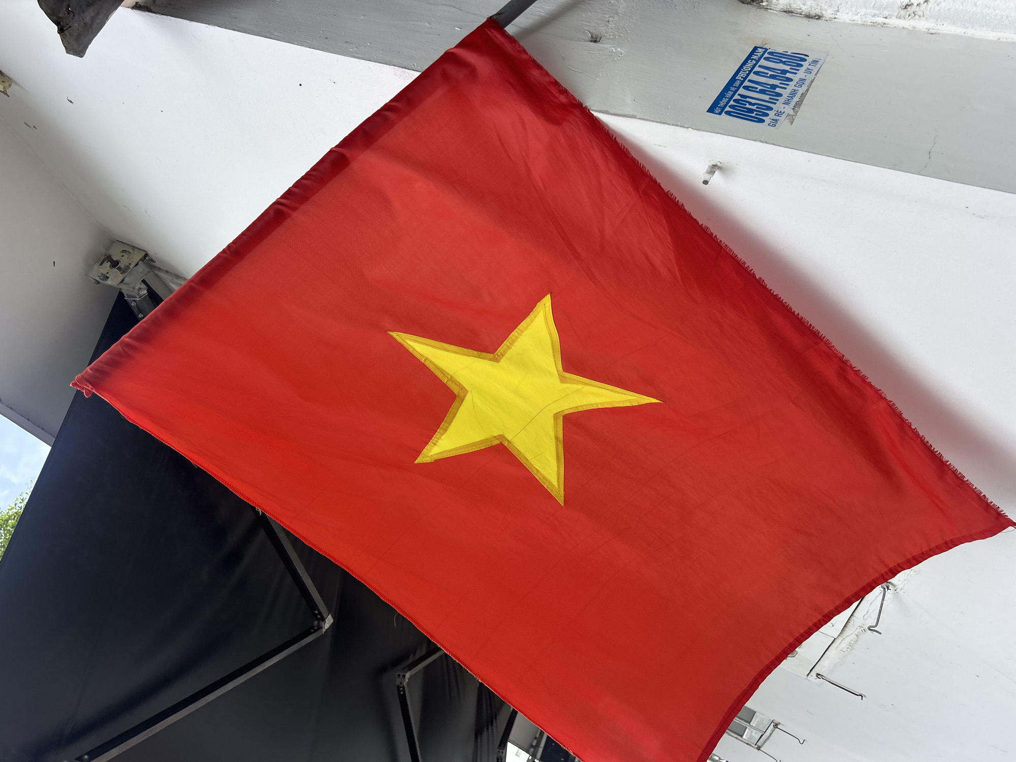 The image depicts the national flag of Vietnam, a red field with a yellow five-pointed star at its center, hanging indoors. The flag is positioned at an angle against a white wall. Above the flag, a blue and white sticker with the phone number 0931.64.64.80 is affixed to the wall, advertising plumbing services in Phuong Nam. The image appears to be taken in Bac Nha Trang Ward, Vietnam. A black canvas awning or similar structure is visible in the lower-left corner, partially obscured by the flag.