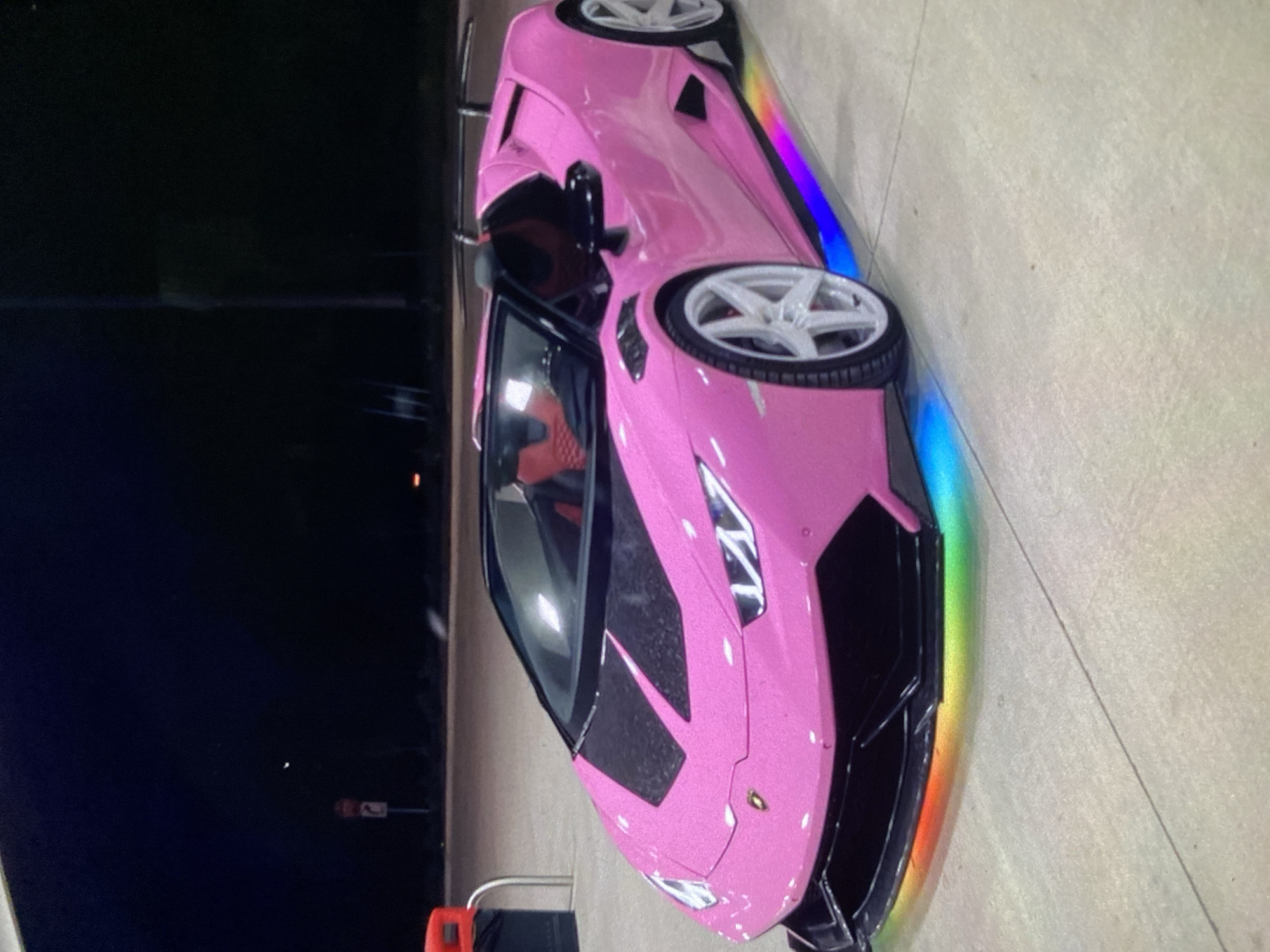 A pink Lamborghini is parked on a concrete surface. The car has a rainbow-colored trim. The background is dark, suggesting it is nighttime. The location and details regarding Lhokseumawe, Indonesia, are not discernible from the image.