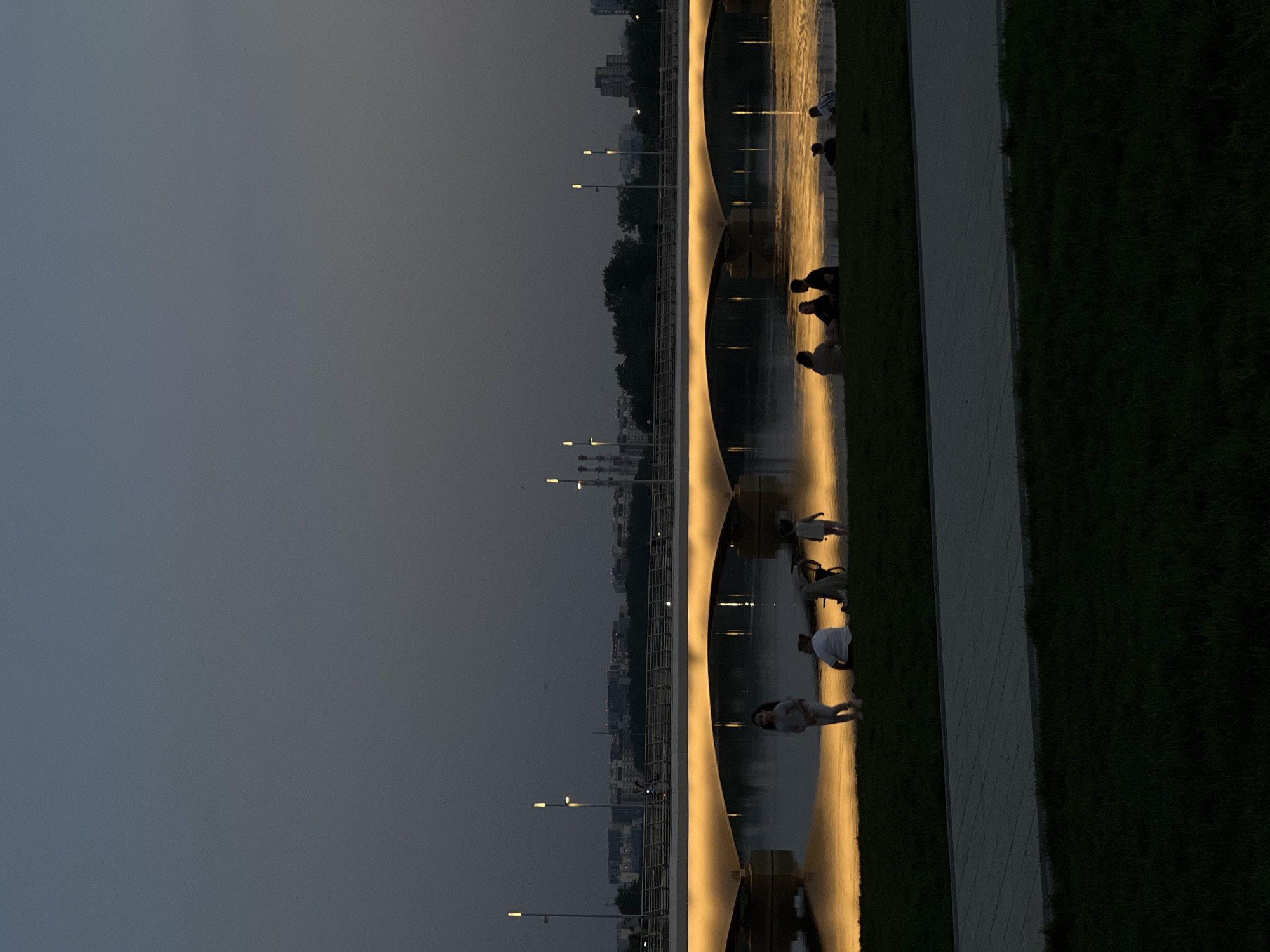 The image captures a view of the Patriarshy Bridge in Moscow, Russia, at dusk or dawn. The bridge structure, illuminated with warm light, is visible, along with the city skyline in the background. Pedestrians can be seen walking and resting along the bridge. A walkway and grassy area border the bridge's edge. The sky above is a uniform grayish tone. The lighting is subdued, creating a reflective effect on the water surface.
