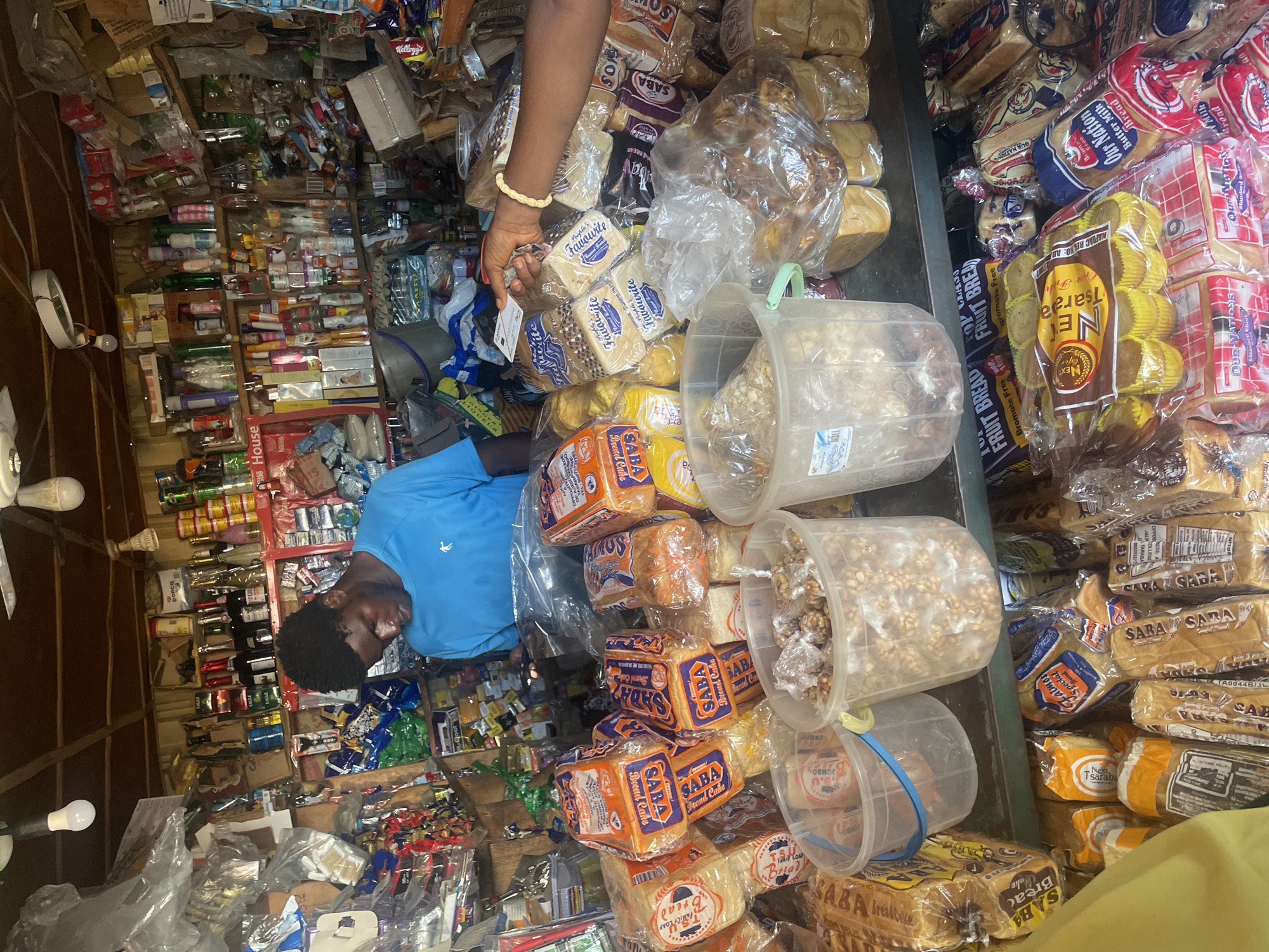 The image depicts a young Black male wearing a blue t-shirt in a crowded convenience store. The store is stocked with a wide variety of goods, including packaged bread, snacks, canned goods, bottled drinks, and toiletries, all arranged on shelves and in bins. The store appears to be located in a humid environment, possibly a market stall or small shop, with open shelving and a ceiling fan visible. In the foreground, there are several loaves of bread packaged in orange and white wrappers, along with containers of nuts and other food items. A hand, likely belonging to another individual, is seen extending a small card or receipt towards the young man. The scene suggests a commercial transaction taking place within a bustling retail environment. The store's contents and arrangement suggest a location in Jalingo, Nigeria, where such small, densely stocked shops are common.