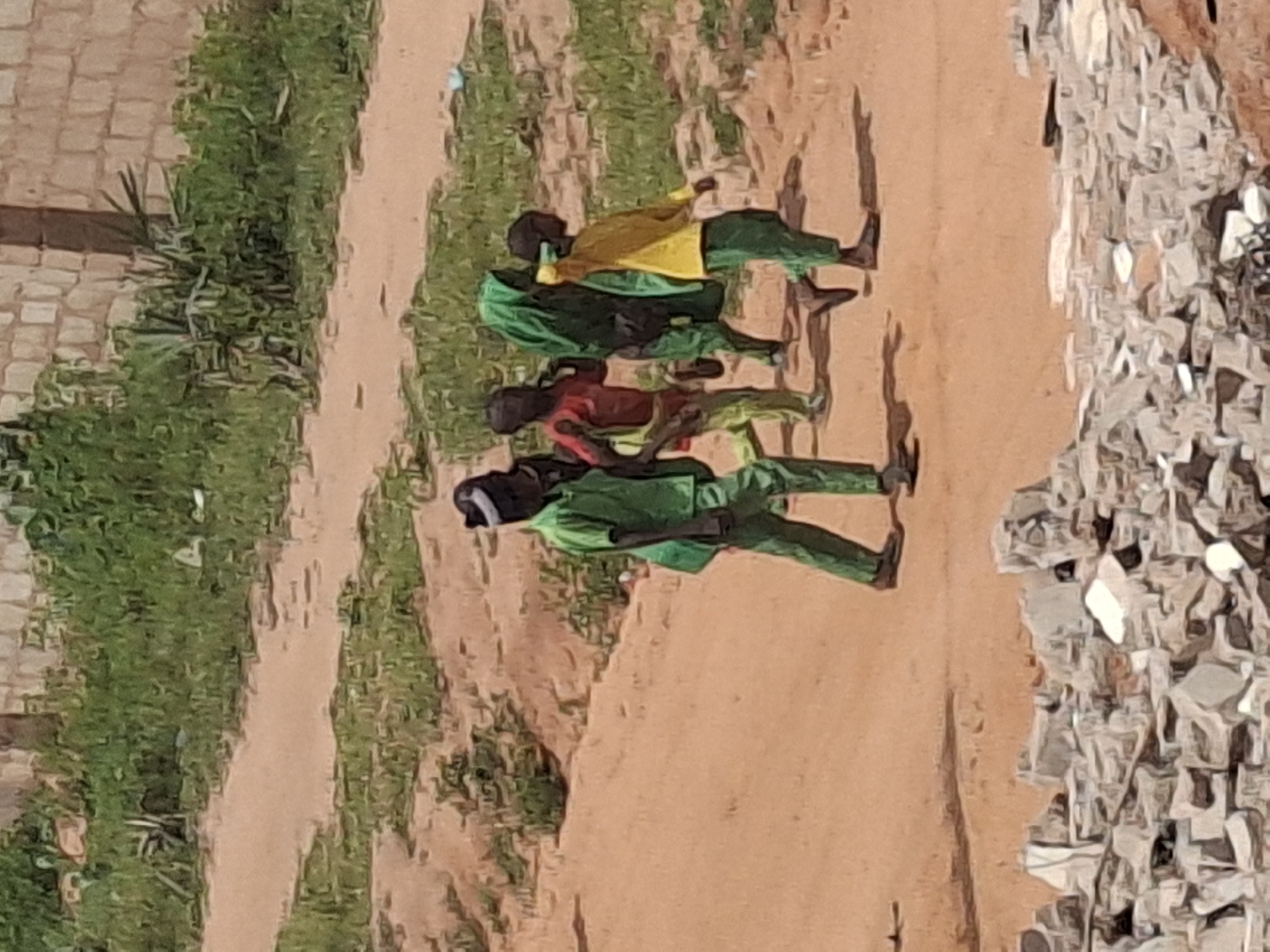 Four children are walking on a dirt path in a sunlit outdoor environment. The child on the far left is wearing a bright green uniform and a black and white headband. To their right, another child in a red shirt and green pants is visible. Next, a child in a green headscarf and green clothing is walking. On the far right, a child in a yellow shirt and green pants is seen walking. In the foreground, there are piles of rubble and debris. In the background, there is green vegetation and a brick wall. The scene appears to be in Tumfure, Nigeria.