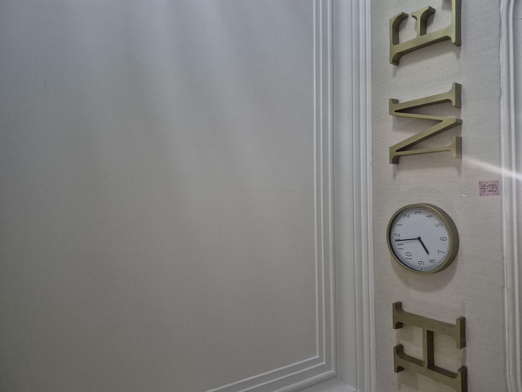 A close-up shot displays a white wall on the left, adjoined by a bordered panel on the right. Gold-colored, block letters spelling "HOME" are vertically aligned on the panel, interspersed with a round wall clock positioned between the "M" and "E". The clock face has black numerals and hands, with the time showing roughly 9:52. The scene is lit softly, highlighting the texture of the wall and the dimensional depth of the letters and clock. The location is inside a home interior in Tbilisi, Georgia.