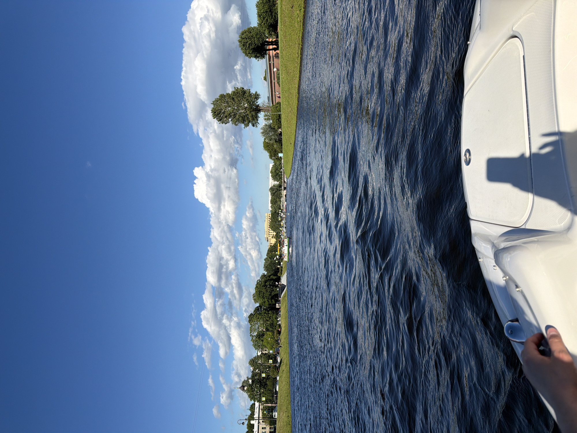 The image shows a view from the front of a boat on a sunny day. The boat is white and appears to be moving through dark blue water with ripples. A person's hand is visible on the left side of the boat. In the background, there is a cityscape with buildings, trees, and a clear blue sky with fluffy white clouds. Several other boats are visible on the water in the distance. The scene suggests a recreational activity on a river or canal within a city.