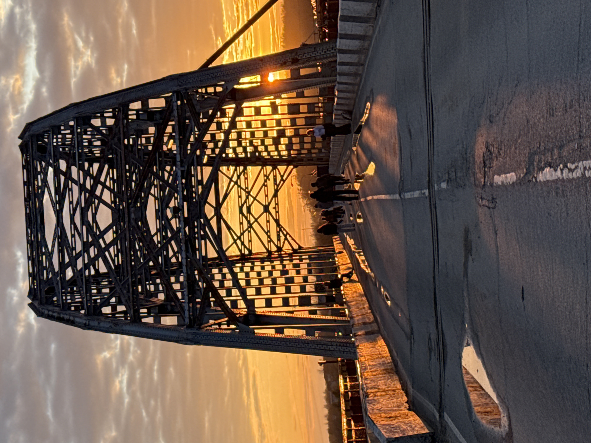 This image captures a picturesque scene of a large metal truss bridge in Dmitrov, Russia, during either sunrise or sunset, characterized by a vibrant golden hour. The sky is illuminated with warm orange and yellow tones, featuring soft, wispy clouds that catch the light.
The prominent subject is the sturdy, dark silhouette of the bridge's metal framework, which dominates the left side of the frame. The sun is visible as a bright glow peeking through the intricate lattice structure of the bridge, creating striking light rays and casting long, defined shadows.
On the paved road surface of the bridge, several individuals are present, mostly appearing as dark silhouettes against the bright backdrop. They seem to be walking or standing, possibly observing the scenic view or enjoying the moment. One person is more clearly discernible, wearing a light shirt and dark pants, standing near the bridge railing. A stone barrier or sidewalk runs along the right side of the road.
Puddles of water are scattered across the road surface, reflecting the intense golden light from the sky, adding a luminous quality to the scene. The overall impression is one of tranquility and natural beauty, enhanced by the dramatic lighting. There is no visible text in the image.