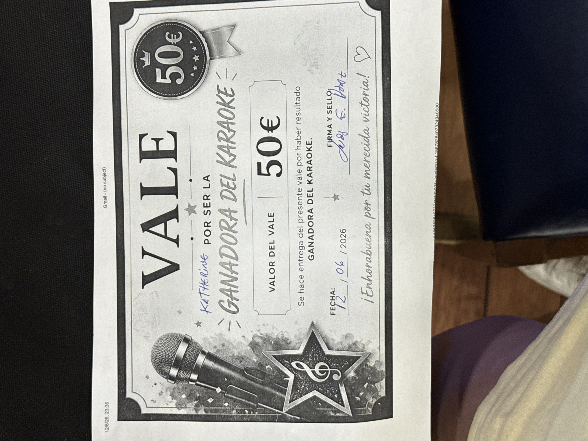 This is a photograph of a certificate or voucher awarding someone named KATHERINE for being the winner of a karaoke event. The certificate has a graphic of a microphone and a star. The text on the certificate reads: "VALE", "KATHERINE POR SER LA GANADORA DEL KARAOKE", "VALOR DEL VALE 50€", "Se hace entrega del presente vale por haber resultado GANADORA DEL KARAOKE.", "FECHA: 12 / 06 / 2026", "FIRMA Y SELLO:", and "¡Enhorabuena por tu merecida victoria!". The certificate is signed with blue ink. The background of the image appears to be a dark, possibly textured surface. The timestamp in the upper left corner indicates "12/6/26, 23:36", and a "Gmail - (no subject)" label is visible at the top. The bottom portion of the image shows the edge of a white paper with some text, and below that, a dark surface which could be a table or floor, with parts of clothing or fabric visible. There is no identifiable information within the image that places the scene in Torrejón de Ardoz, Spain.